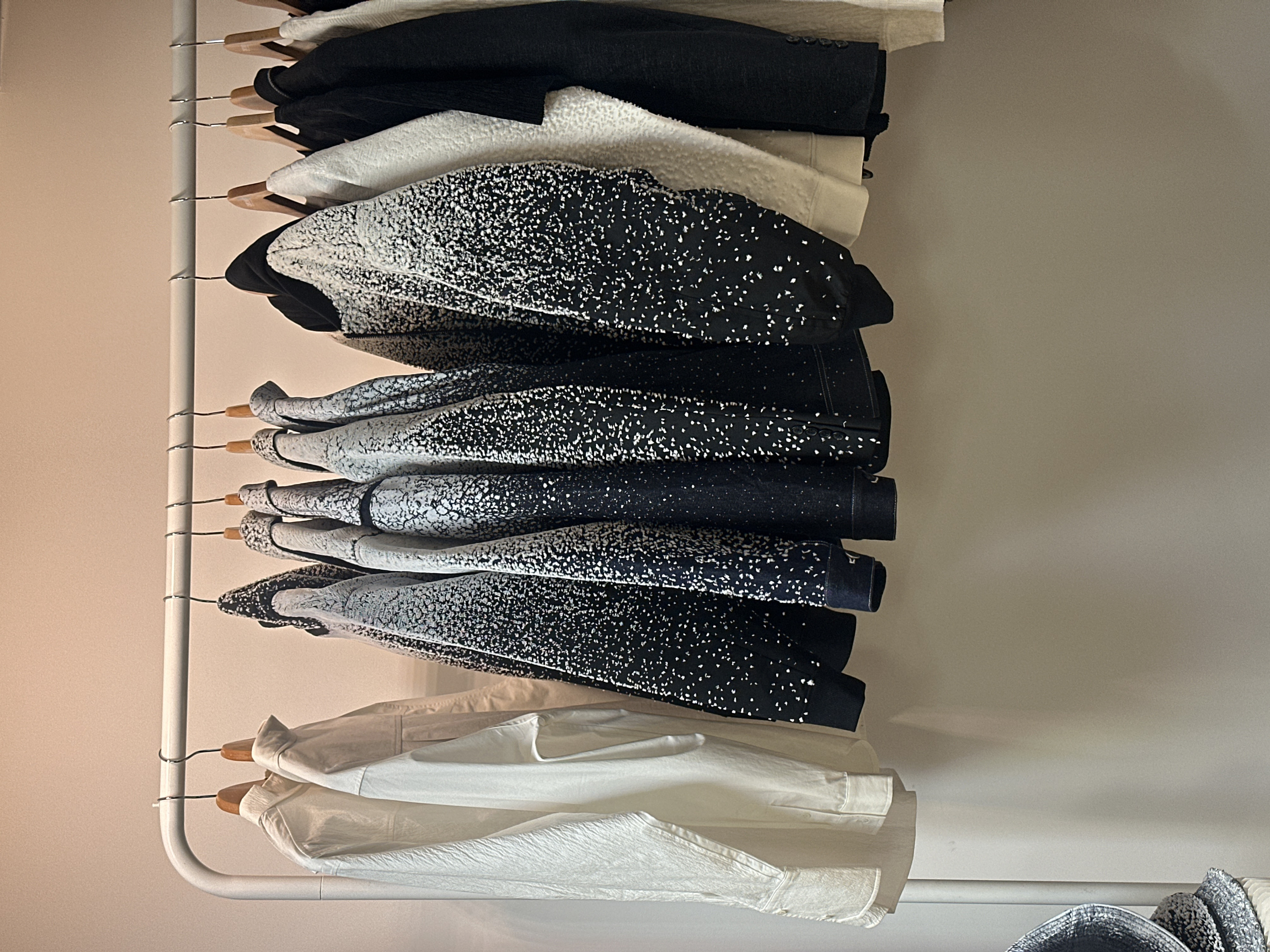 The image presents a close-up view of various garments neatly organized on a white metal clothes rack, positioned vertically against a plain, light-colored wall. The clothes are stacked one below the other, creating a layered effect of textures and patterns.
At the top of the stack, a dark, likely black, garment resembling a blazer or jacket is partially visible, followed by a cream or off-white piece with a soft, possibly fuzzy or sherpa-like texture. Immediately beneath this, a prominent pattern emerges: several garments, which appear to be jackets or long-sleeved tops, feature a striking black base with an all-over white speckled or dotted design. This pattern varies in density, appearing more concentrated and white-dominated in some areas, and sparser and black-dominated in others, suggesting a gradient or unique artistic print.
Further down the rack, more items continue this distinctive black and white speckled pattern, with some showing sleeves or cuff details. The texture of these speckled garments seems slightly coarse due to the print. Below these, the collection transitions to lighter-colored items. A stack of white or off-white shirts or blouses is visible, with the topmost appearing to be a smooth, classic collared shirt. The garment directly below it showcases a distinct crinkled or textured fabric, adding another tactile element to the composition.
The clothes are hung on a combination of wooden hangers with metal hooks and simpler metal wire hangers, all suspended from the white metal clothes rack. The rack itself is composed of a vertical bar with horizontal extensions, providing a sturdy support structure. The background wall is unadorned and uniform in color, likely a light beige or off-white, allowing the focus to remain entirely on the clothing. There are no people present, and no text is visible within the frame. The lighting appears soft and diffused, highlighting the different textures and the monochromatic palette of the garments.