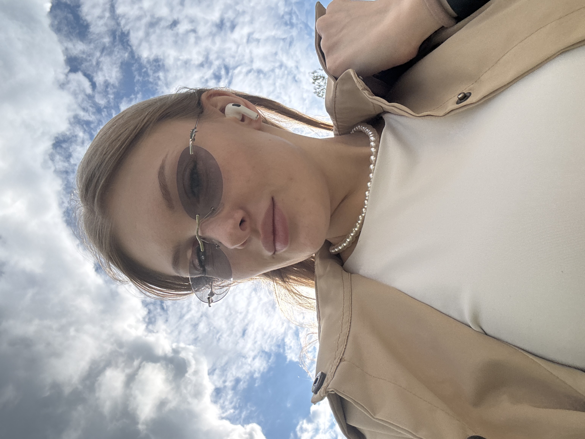 A young woman is depicted in a close-up, low-angle portrait, looking upwards towards the camera. The background is a bright blue sky filled with white, fluffy cumulus clouds, suggesting an outdoor setting during the daytime.
The woman has light-colored hair, styled loosely around her face. She is wearing light-tinted, oval-shaped sunglasses with thin metal frames. A white Apple AirPod earbud is visible in her right ear. Around her neck, she wears a simple pearl necklace. Her attire consists of a plain white top, partially covered by a light beige or tan jacket or coat, which features a visible metal snap button on the lapel area. Her facial expression is neutral and composed. There is no visible text in the image.