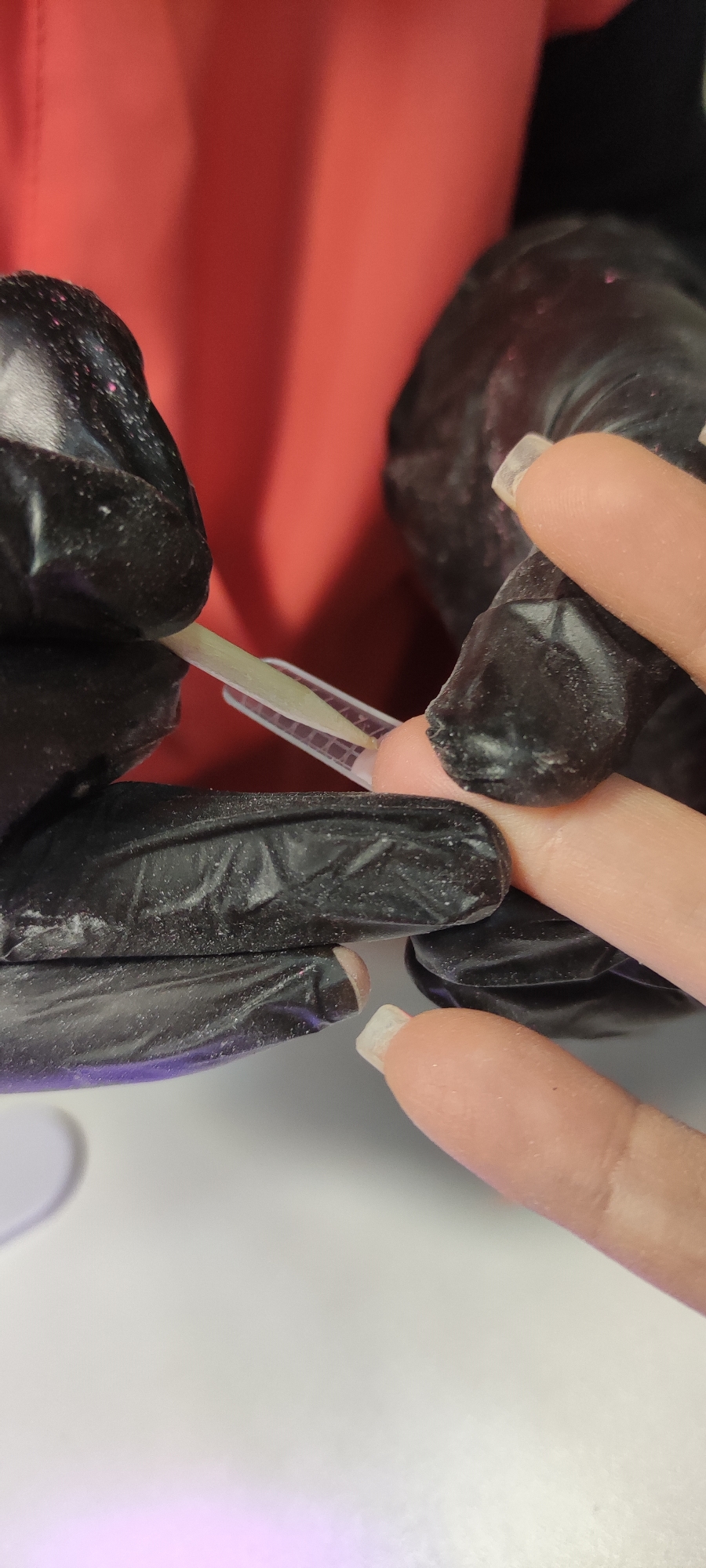 A close-up shot depicts a person in black gloves applying a nail form to another person's fingernail. The nail form is clear with a grid pattern. The person receiving the nail application has fair skin and short, natural nails. The background includes a white surface and a reddish-orange garment. There's no information in the picture that indicates the location is in Chaikovsky, Russia.
