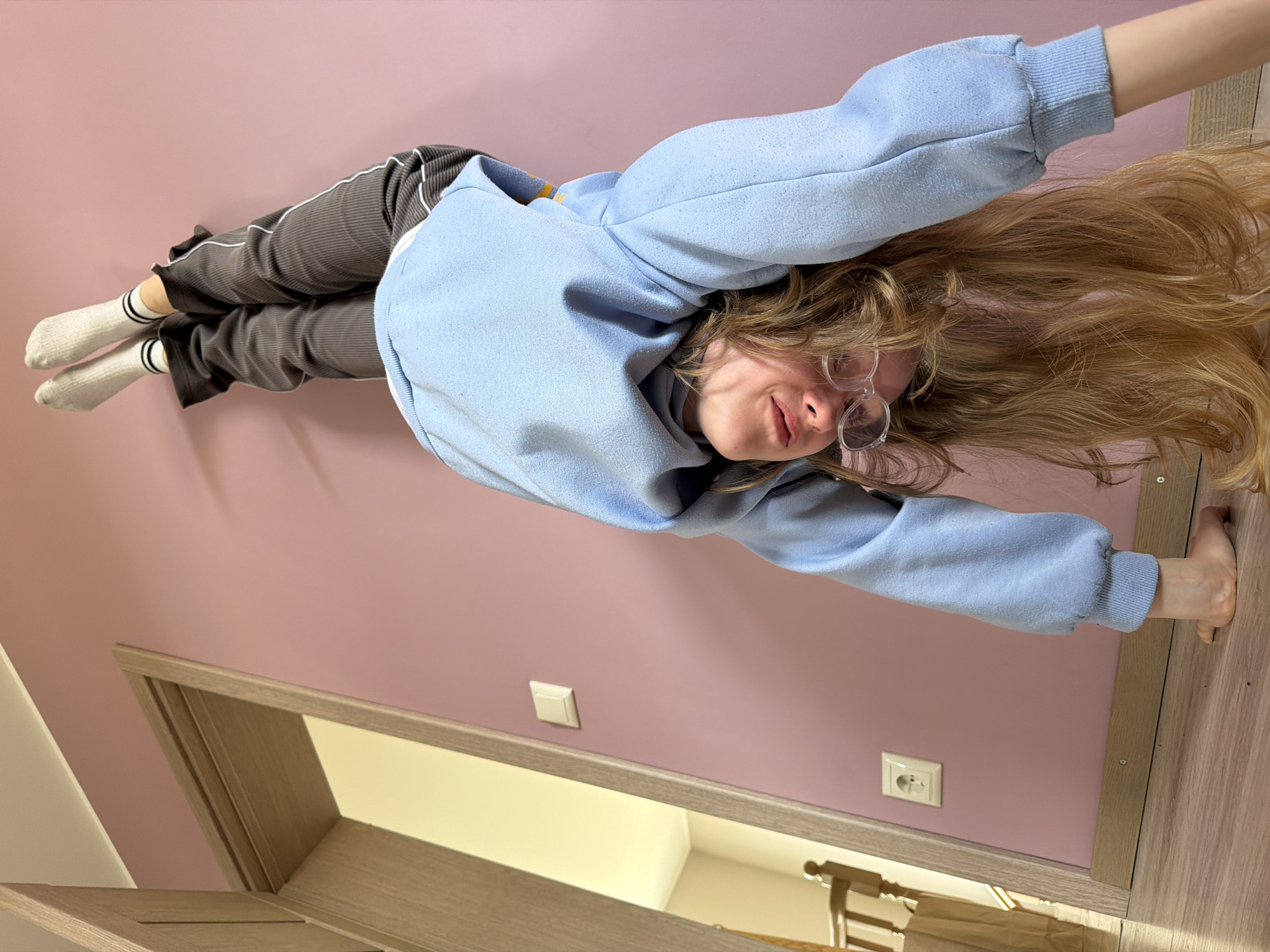 A young woman is standing on her hands against a pink wall in what appears to be an indoor space. She is wearing a light blue hooded sweatshirt, gray ribbed pants with white stripes, and white socks with black stripes. Her long, light brown hair is hanging down. She is wearing glasses. A light switch and an electrical outlet are visible on the wall. A partially open wooden door is in the lower left corner, revealing a glimpse of another room with what appears to be furniture or items stored inside. The location is indicated as Dreiliņi, Latvia, though no location-specific details confirm this.