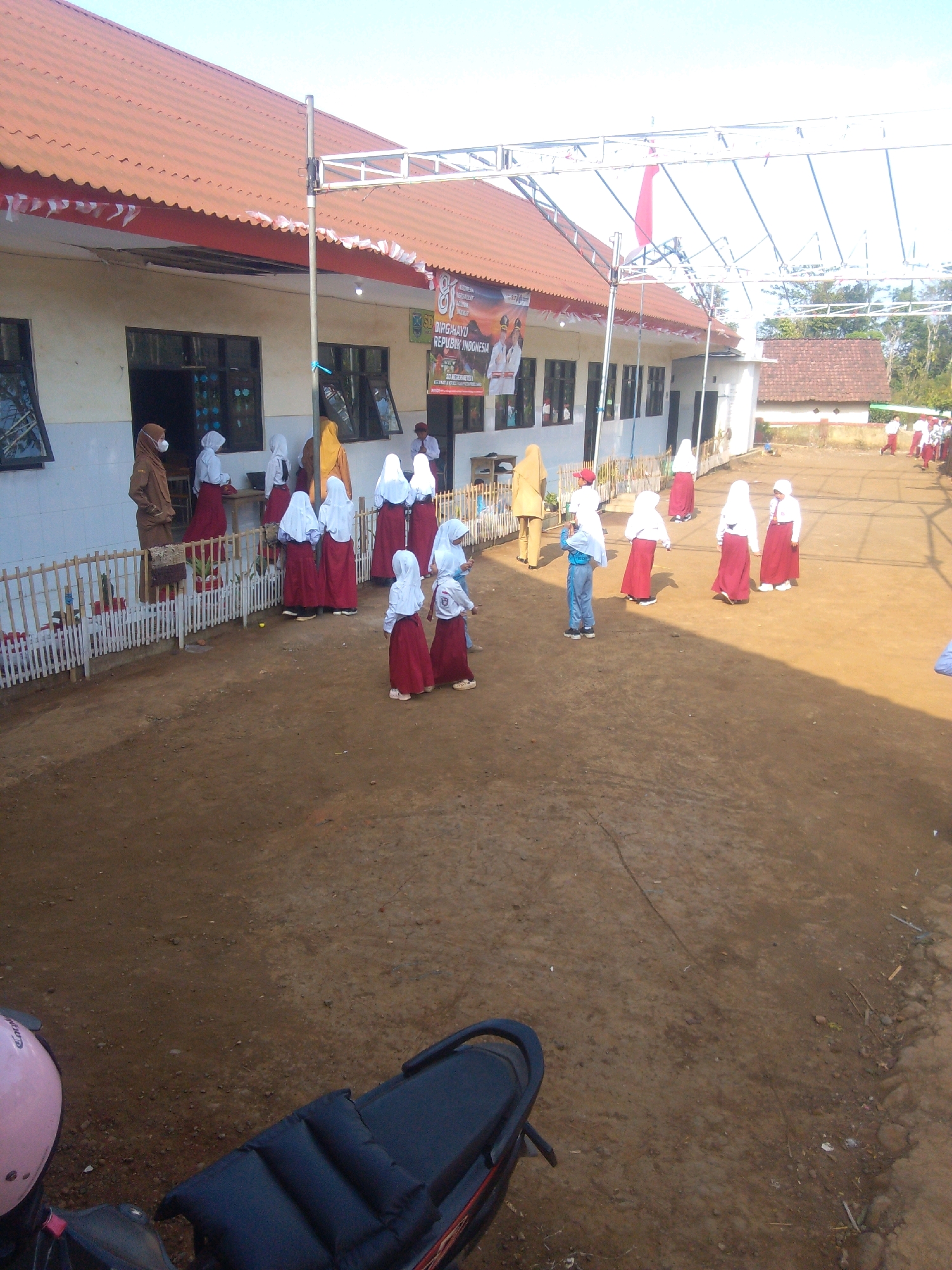 The image depicts an outdoor scene at what appears to be a school in Betek, Indonesia. In the foreground, the rear of a dark-colored motorcycle with a pink helmet is visible. The ground is predominantly dirt. The main focus is on a single-story building with a reddish-orange tiled roof and white walls. Several windows line the building.
In front of the building, a group of individuals, primarily young girls in white uniforms with red skirts and headscarves, are scattered across the dirt area. Some are standing in small groups, while others are more isolated. One adult woman in a brown hijab and a tan jacket stands near the building. Another adult, wearing a white shirt and a dark tie, is seen near the entrance of the building. A banner with Indonesian text, likely commemorating Indonesian Independence Day, is displayed on the building. A temporary metal structure with flags or banners is erected over the area in front of the building. The overall environment suggests a sunny day with clear skies.