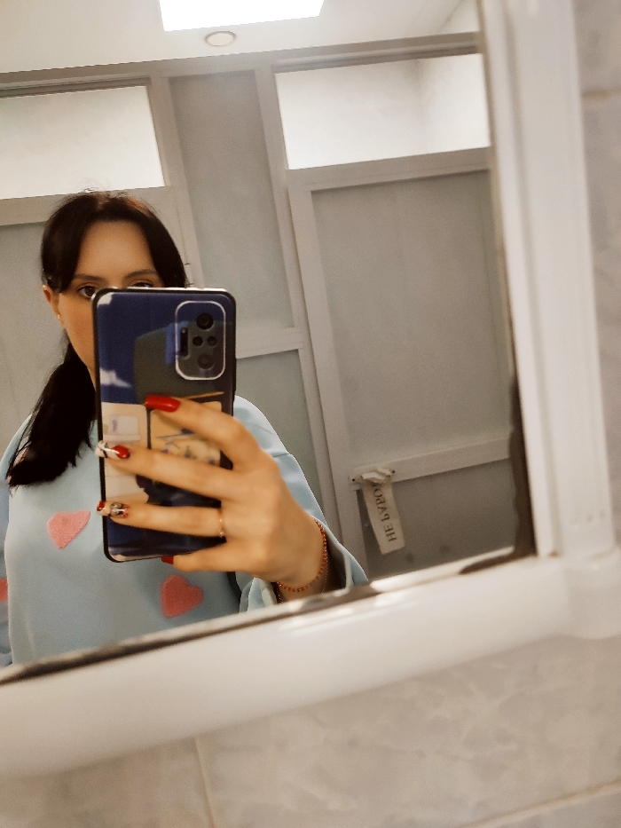 The image depicts a female individual taking a mirror selfie in an indoor setting. The specific location within the city of Donetsk, Ukraine, cannot be determined from the visual information provided.
The individual, seen from the upper torso, has dark hair tied back, with strands framing her face. Her right eye and the bridge of her nose are visible; the rest of her face is obscured by a dark blue smartphone, held in her right hand. She is wearing a light blue long-sleeved top adorned with multiple pink, heart-shaped appliques. Her right hand displays long fingernails painted red, with some featuring a white and black pattern. A ring is present on her right ring finger, and a thin, reddish-orange bracelet is visible on her right wrist. The smartphone case is dark blue, possibly with a light blue and white cloud-like design on the lower portion, and its multi-lens camera module is visible.
The environment, reflected in the mirror, appears to be a tiled room, possibly a restroom or changing room, with light-colored, possibly off-white, tiled walls. Above the mirror, a white square light fixture with a circular central light is installed on the ceiling. The background features light-colored walls or partitions, with horizontal frosted or opaque panels at the top, consistent with cubicle-style construction. A light-colored fabric tag or strap, hanging from a fixture on the wall to the right of the subject's reflection, displays Cyrillic text, partially legible as "НЕ БУРО..."