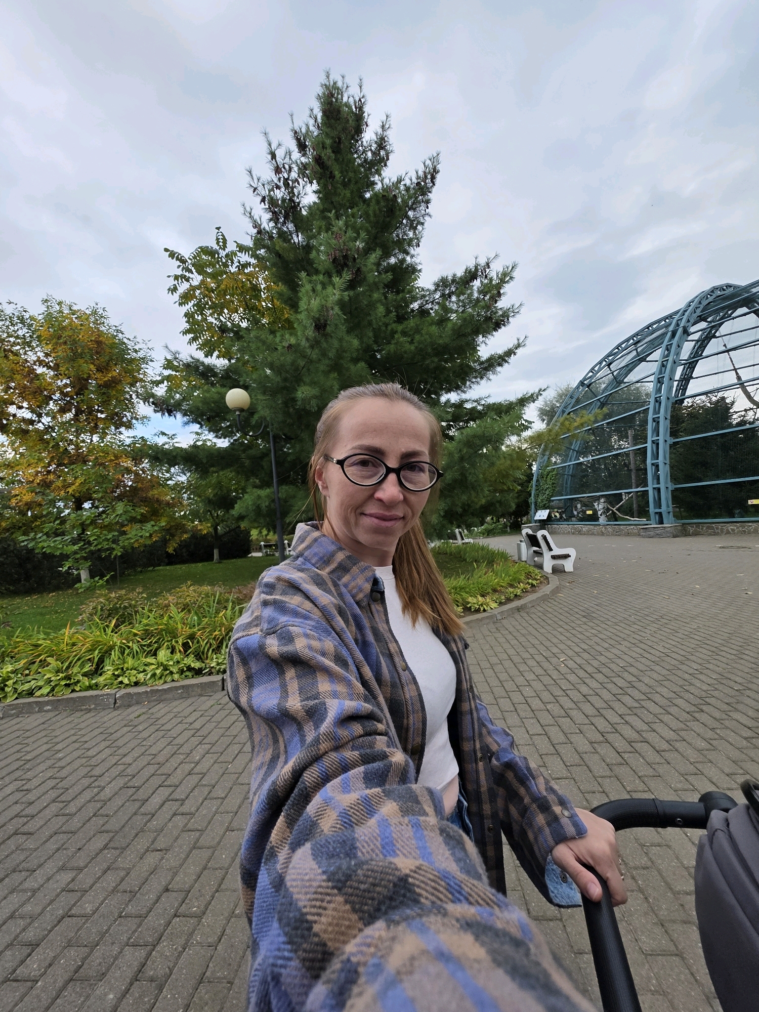 The image shows a woman with blonde hair and glasses, wearing a plaid shirt and a white top, pushing a stroller on a paved path in a park. Behind her, there is a large evergreen tree, a street lamp, and lush green foliage. To the right, a large, arched aviary with blue metal framing is visible, with several birds inside. The aviary appears to be part of a zoo or wildlife park. The overall environment suggests an outdoor recreational area within a city, possibly the Udmurt State Zoo in Izhevsk, Russia, given the aviary's design and the presence of park-like surroundings. The sky is overcast, indicating a cloudy day.