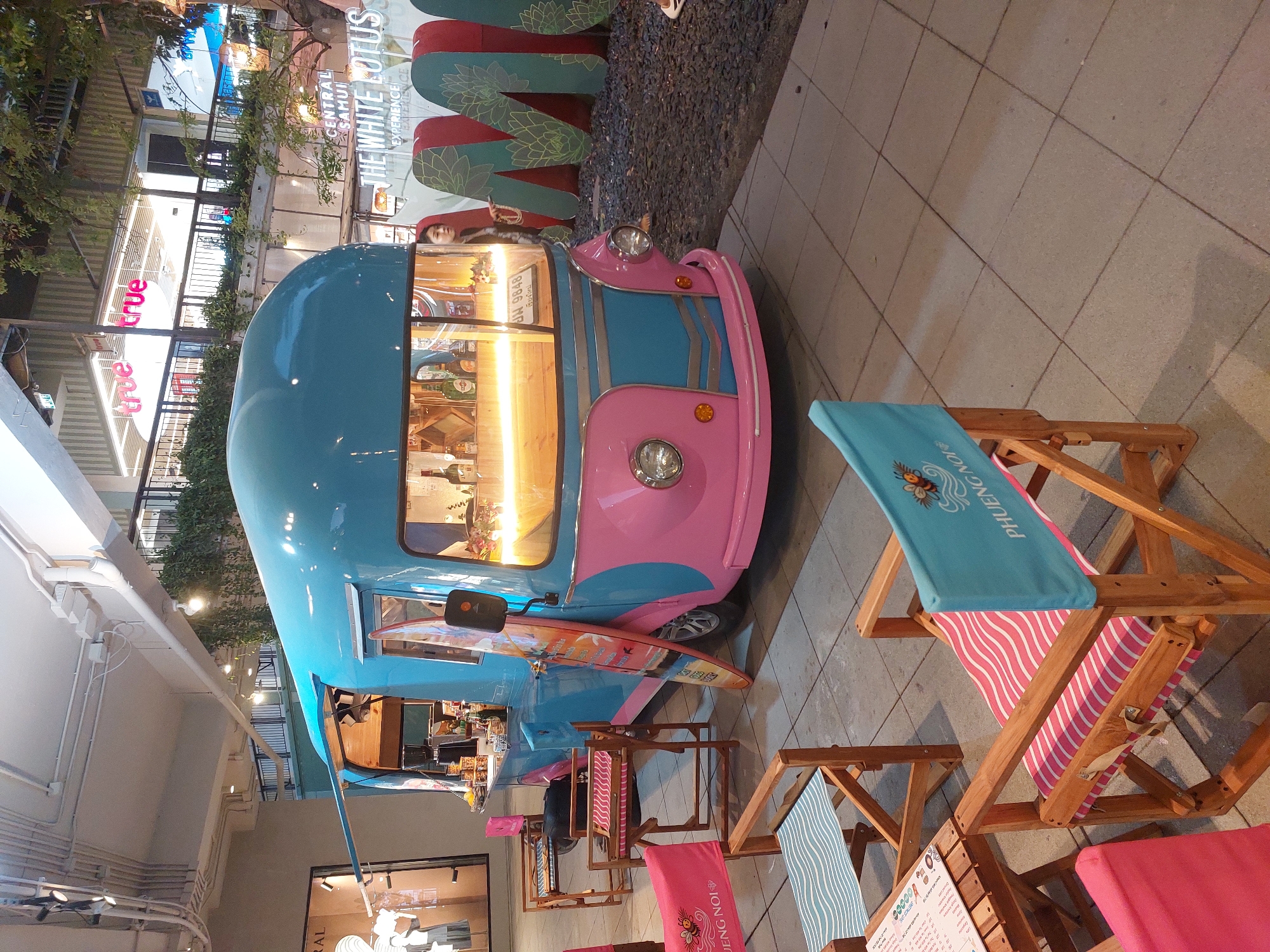 In Baan Chaweng, Thailand, a vibrant blue and pink food truck with a retro design is parked on a paved outdoor plaza. The food truck is illuminated from within, showcasing bottles of liquor and a coffee machine on the counter. Adjacent to the truck, several wooden director's chairs with colorful striped cushions and canvases are set up, suggesting an outdoor seating area. One chair prominently displays the text "PHUENG NOI" and an image of a bee. In the background, large, decorative green letters spell out "M," with other letters partially visible, and a building with "true true" signage is visible, indicating a commercial area. The scene appears to be an evening setting, with artificial lighting contributing to the atmosphere.