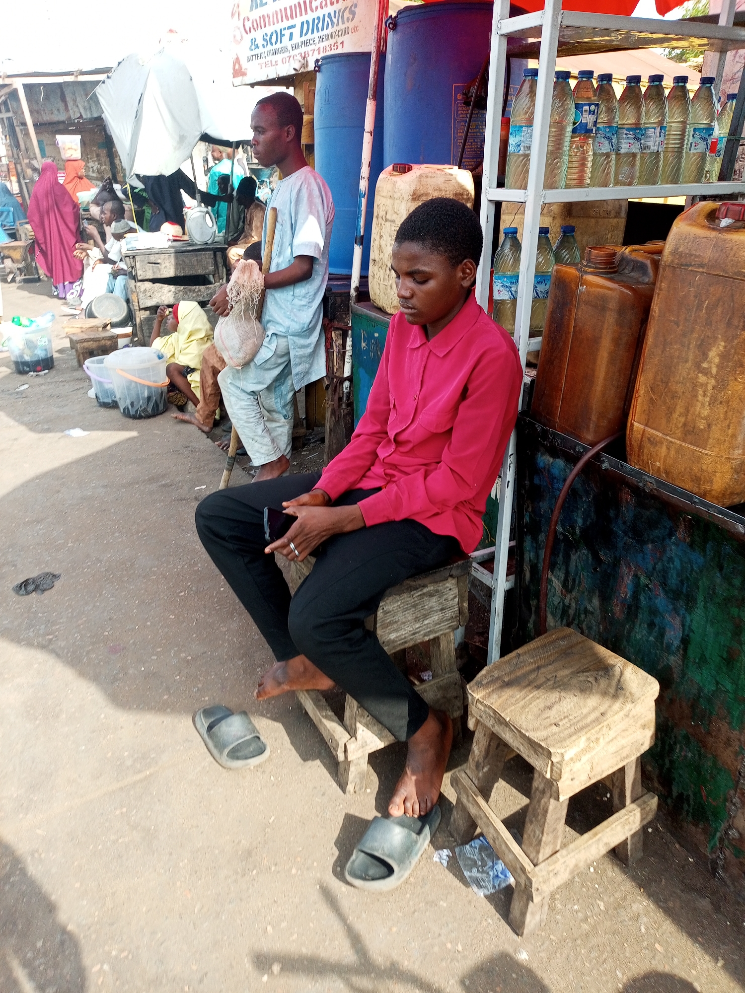 Here's a detailed description of the image:
**Setting:** An outdoor scene in an unidentified area of the city. The environment appears to be a bustling marketplace or roadside business area.
**People:**
* **Central Figure:** A young person is sitting on a small wooden stool, looking down at their mobile phone. They are wearing a red button-down shirt and black pants. They are barefoot.
* **Standing Man:** A man stands in the background, wearing a light-colored, long-sleeved outfit. He is holding a stringy gourd.
* **Other Individuals:** There are several other people in the background, some sitting and some walking. One individual is wearing a distinctive red robe.
**Objects:**
* **Stools:** There are two small, simple wooden stools.
* **Mobile Phone:** The seated person is holding a mobile phone.
* **Containers:** Several large plastic containers are visible, including large blue barrels and brown jerrycans.
* **Water Bottles:** Numerous bottles of water are displayed on a metal rack.
* **Signage:** A sign is present, displaying information for "Communications & Soft Drinks."
* **Sandals/Flip-flops:** A pair of sandals/flip-flops are laying on the ground.
**Action & Interactions:**
* The central person is engaged in using their mobile phone, possibly texting or browsing.
* The man with the gourd is simply standing there.
* The environment suggests a place of commerce and activity, where people are either buying, selling, or passing through.
**Overall Impression:** The image captures a moment in a commonplace setting, likely a market or roadside stall in an urban area. The focus seems to be on the seated individual and their use of technology within this environment.