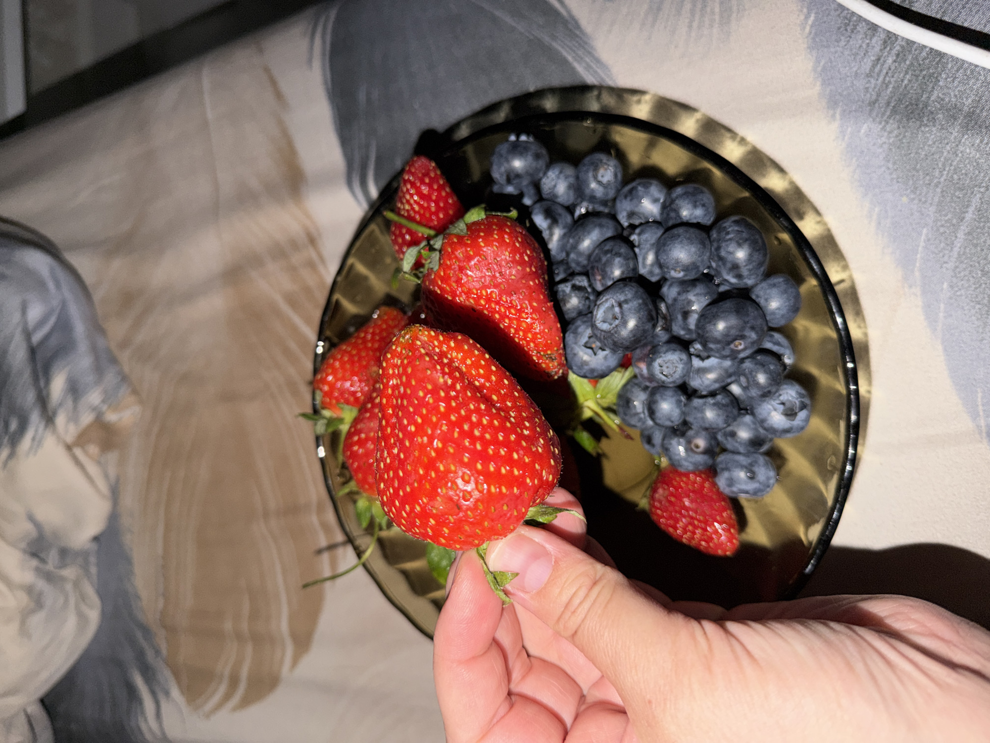 A top-down shot shows a clear brown glass bowl containing fresh blueberries and strawberries. A hand holds a strawberry above the bowl. The background features a textured fabric surface. The location or city is not determinable from the image.