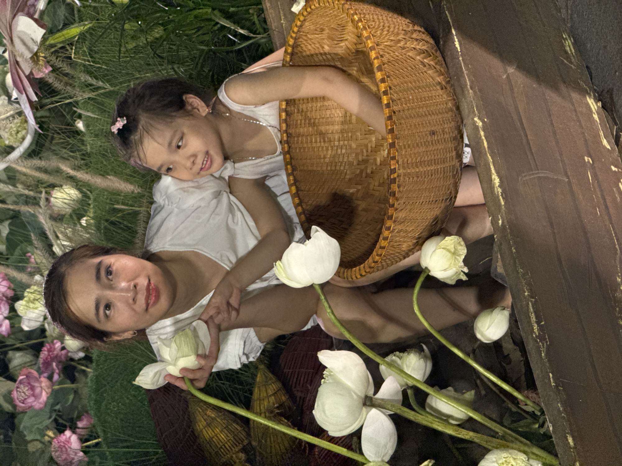 The media file depicts two individuals, an adult and a child, in an outdoor setting surrounded by greenery and white lotus flowers. The adult, a woman with dark hair, is looking towards the camera with a gentle smile, holding a lotus flower. She is wearing a white top. The child, a young girl with dark hair adorned with a pink clip, is lying on the woman's shoulder and looking away from the camera. She is also wearing a white top.
They are positioned next to a woven basket, which appears to be empty. The scene is filled with numerous white lotus flowers and some pink flowers, suggesting a natural or garden-like environment. The background consists of lush green foliage and what appears to be a wooden structure, possibly a bench or railing. The lighting suggests it might be evening or a dimly lit area.
The overall mood of the image is peaceful and tender, with the interaction between the woman and child conveying warmth and affection. The presence of lotus flowers evokes a sense of tranquility and potentially cultural significance, given the implied location of Hanoi, Vietnam, where lotus flowers are prominent. There is no visible text in the image.