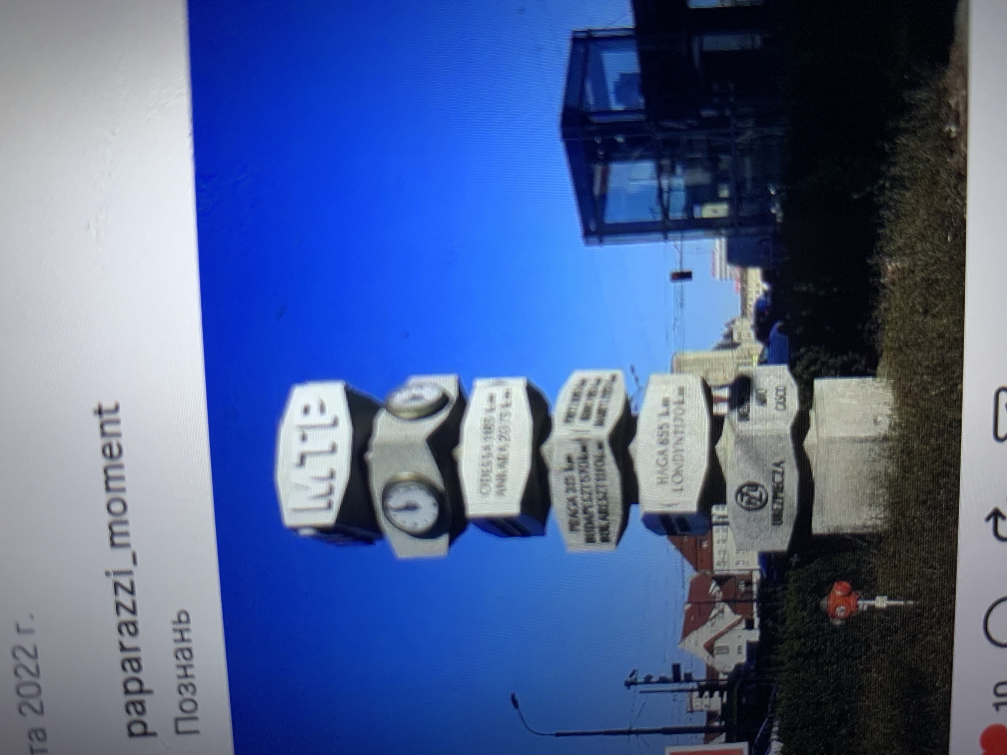 The image shows a tall, multi-sided signpost with various destinations and distances listed on its panels. The top panel displays "MTP". Below this are two clock faces. Further down, there are panels with text such as "ODESSA 1185 km", "ANKARA 2075 km", "PRAGA 315 km", "BUDAPEST 570 km", "BUKAREST 1110 km", "HAGA 855 km", and "LONDYN 1170 km". One panel features a circular logo with the number "77" inside it and the text "GRZĘZĘCZA" below. Another panel has "OSCO". The signpost is located in an outdoor setting with a bright blue sky. In the background, there are buildings, a street light, a fire hydrant, and a modern glass and dark-framed structure. The scene is indicative of a public space within a city.