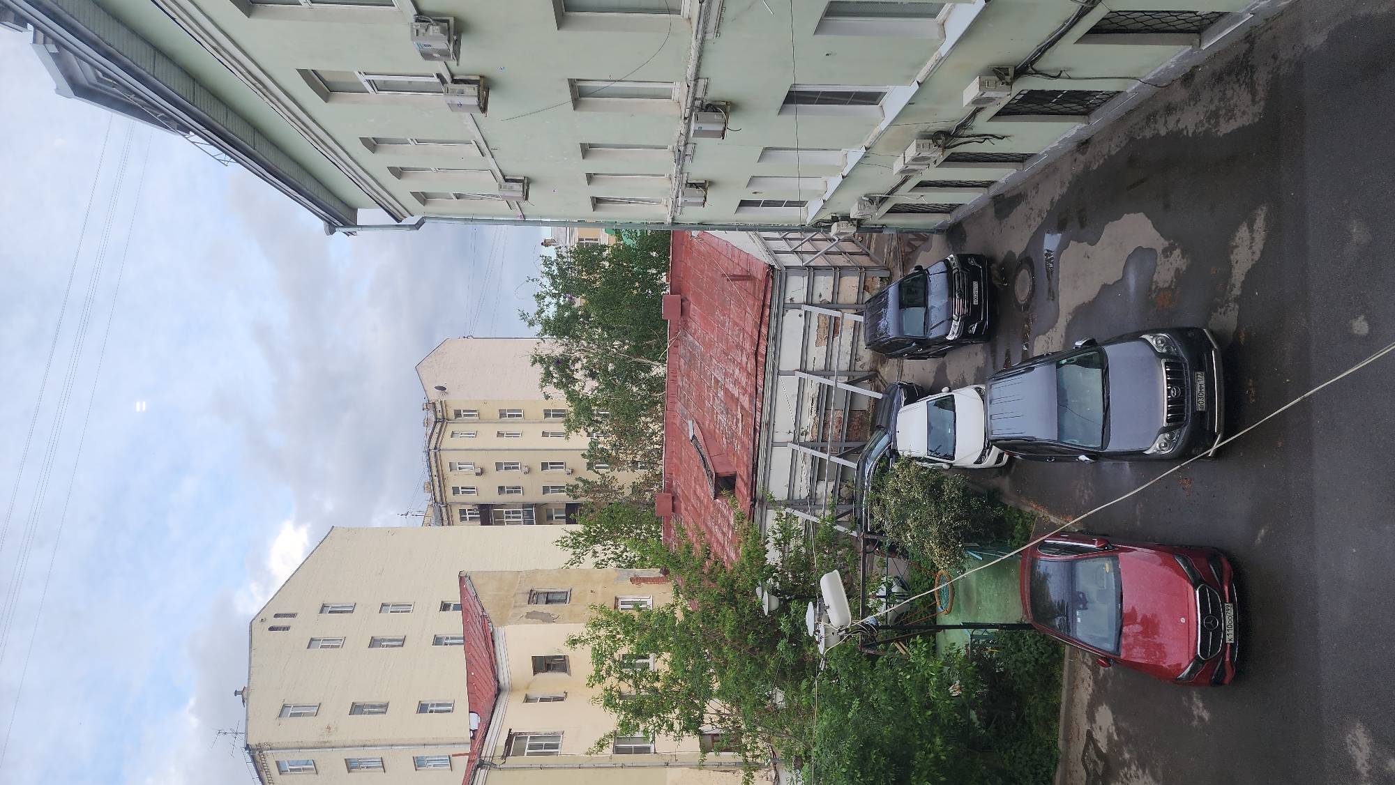 An aerial view of a courtyard in Moscow, Russia. Several cars of different colors are parked. A red-roofed building is in the center. Buildings are on either side of the courtyard. The sky is cloudy.