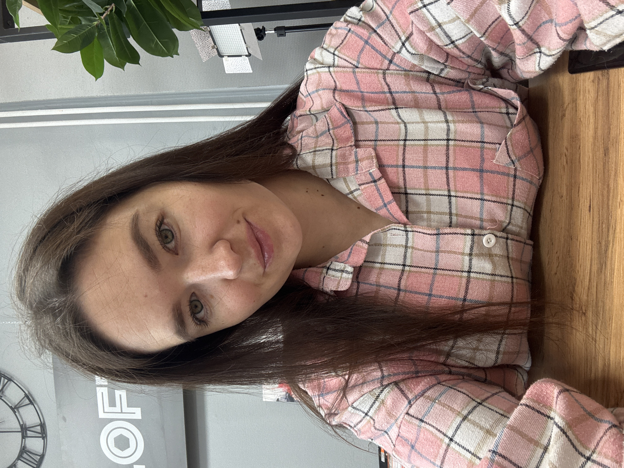 The image, originally oriented 90 degrees clockwise, captures a young woman indoors in what appears to be Chaikovsky, Russia. She has long, dark brown hair and a fair complexion with light freckles across her nose and cheeks. Her eyes are green or hazel, and she has a soft, neutral expression with a slight, gentle smile. She is wearing a pink, white, blue, and black plaid button-up shirt.
The background features a contemporary interior setting. To the upper left, green leaves from a plant are visible. Above and behind her right shoulder, a white light panel or fixture is present. The wall behind her is a light grey, and an industrial-style clock with Roman numerals is partially visible to her lower left. On the grey surface below the clock, the partial text "LOFT" is visible in white letters, hinting at the establishment's name or style. A warm-toned wooden surface, likely a table or desk, occupies the right side of the frame. The woman is positioned for a close-up shot, possibly a selfie, with her head tilted.