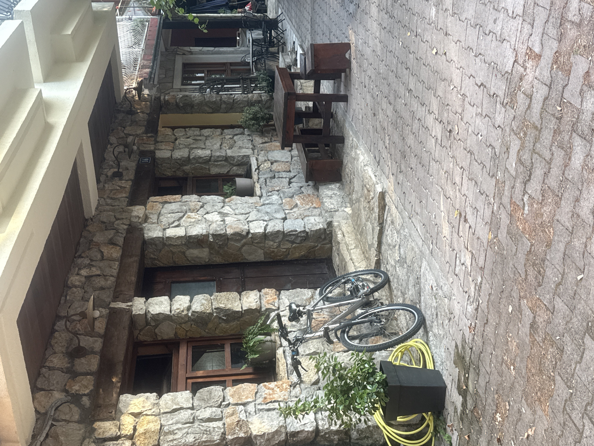 The image depicts the exterior of a building in Virpazar, Montenegro, constructed with rough-hewn stone. In the foreground, a paved walkway, made of interlocking brick pavers, shows signs of moisture and scattered debris. A black planter with a green shrub and a yellow coiled hose are positioned to the left. A silver bicycle leans against the stone wall. Further into the scene, a dark wooden table is accompanied by two wooden benches or stools. Beyond this, outdoor seating with tables and chairs is visible, suggesting a cafe or restaurant setting. The building features wooden-framed windows and doors, and several exterior lights are mounted on the stone facade.