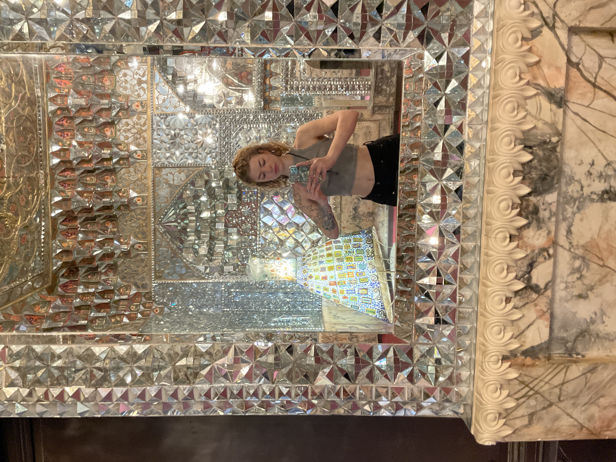 A young woman with curly, reddish-blonde hair is captured in a mirror selfie, holding her phone to photograph her reflection. She wears a light gray crop top and dark pants. Visible tattoos adorn her left arm, featuring floral or organic designs. Her expression is calm and focused on her phone.
The setting is an elaborately decorated interior, likely in Tbilisi, Georgia, characterized by extensive mirror mosaic artwork known as "Aina-Kari" or Shiraz Glasswork. The large mirror reflecting the woman is composed of countless small, faceted mirror pieces arranged in intricate geometric and kaleidoscopic patterns. Some sections of the mirror mosaic also incorporate gilded or painted floral and abstract motifs behind the glass.
The outer frame of the larger mirror features a thick, ornate border with a carved or molded design of scrolls and foliate elements, possibly made of plaster or stone, and appears to be in a light, natural stone or marble-like finish. Beyond this frame, to the right, a wall with a marbled pattern is visible.
The mirrored walls of the room create multiple reflections, showcasing the depth and complexity of the mosaic work. Reflected within the main mirror, behind the woman, is a colorful, possibly modern, light fixture or art installation with a checkered or segmented pattern, contributing to the vibrant and elaborate ambiance. No discernible text is visible in the image.