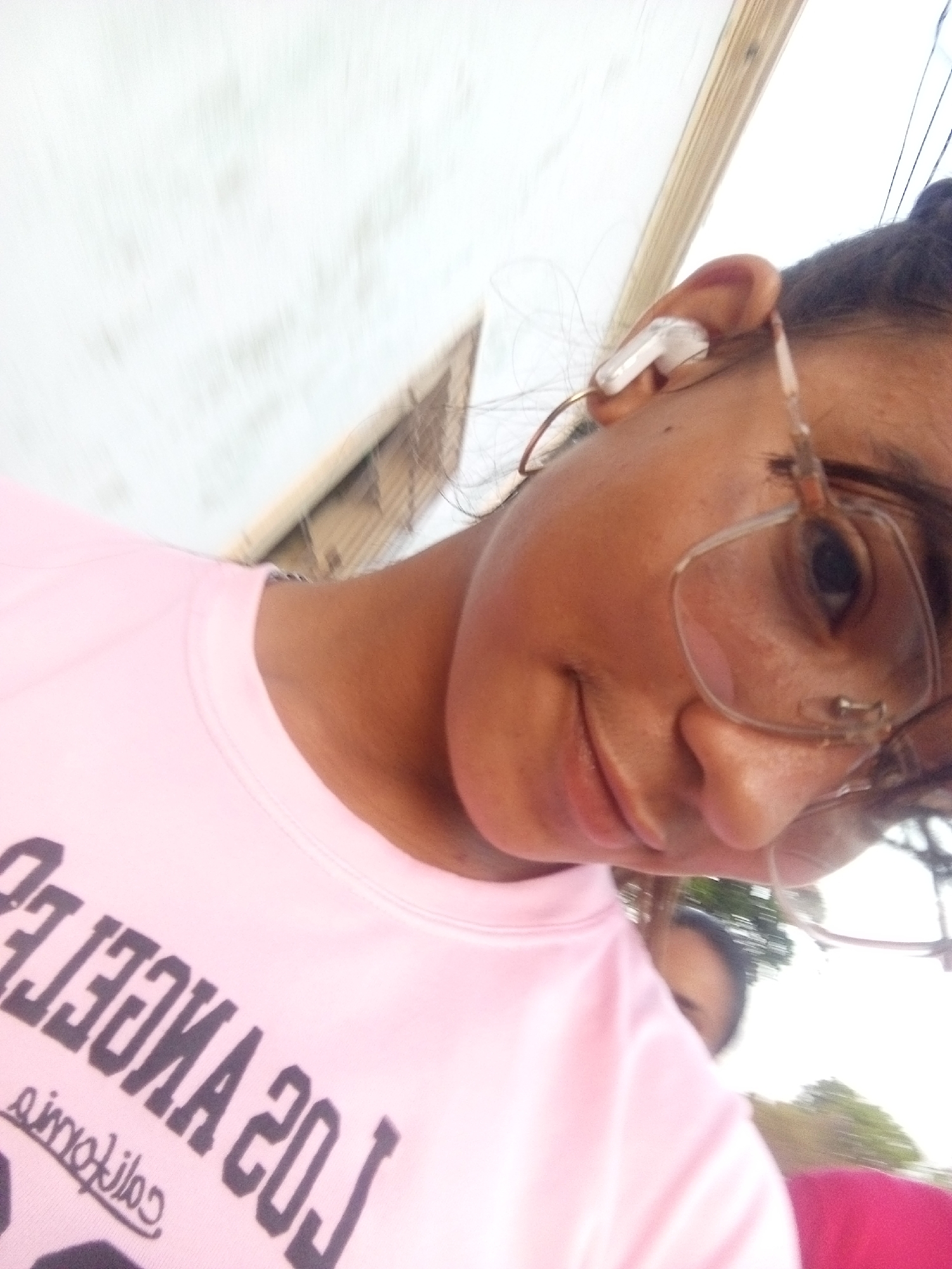 A close-up, rotated selfie shows a person with brown skin wearing glasses and a white wireless earbud in their right ear. They are wearing a light pink t-shirt with the words "LOS ANGELES" printed in reverse. The location is indicated as Juan Griego, Venezuela, but the image does not provide visual confirmation of this location. Another person is partially visible behind the first person, and some foliage can be seen in the background. A white structure, possibly a building or roof, is visible in the upper left corner of the image.