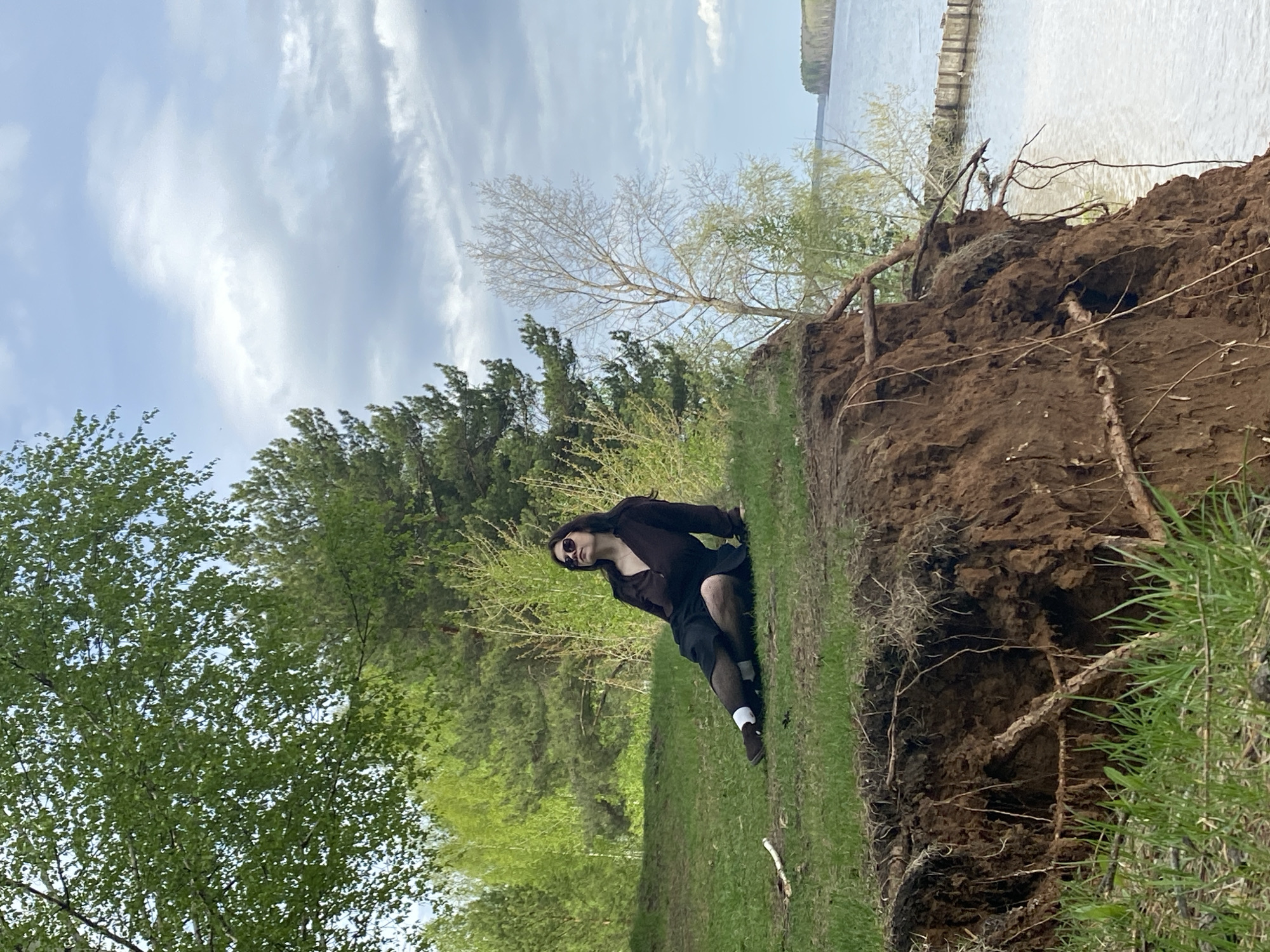 The image shows a person standing on a grassy slope next to a dirt embankment. The person is wearing a dark top, a dark skirt or shorts, and dark stockings or socks with white trim. Sunglasses are worn. In the background, there are trees and a body of water, possibly a river or lake. The sky is partly cloudy. The location is the city of Chaikovsky, Russia. The foreground shows disturbed soil and exposed roots.