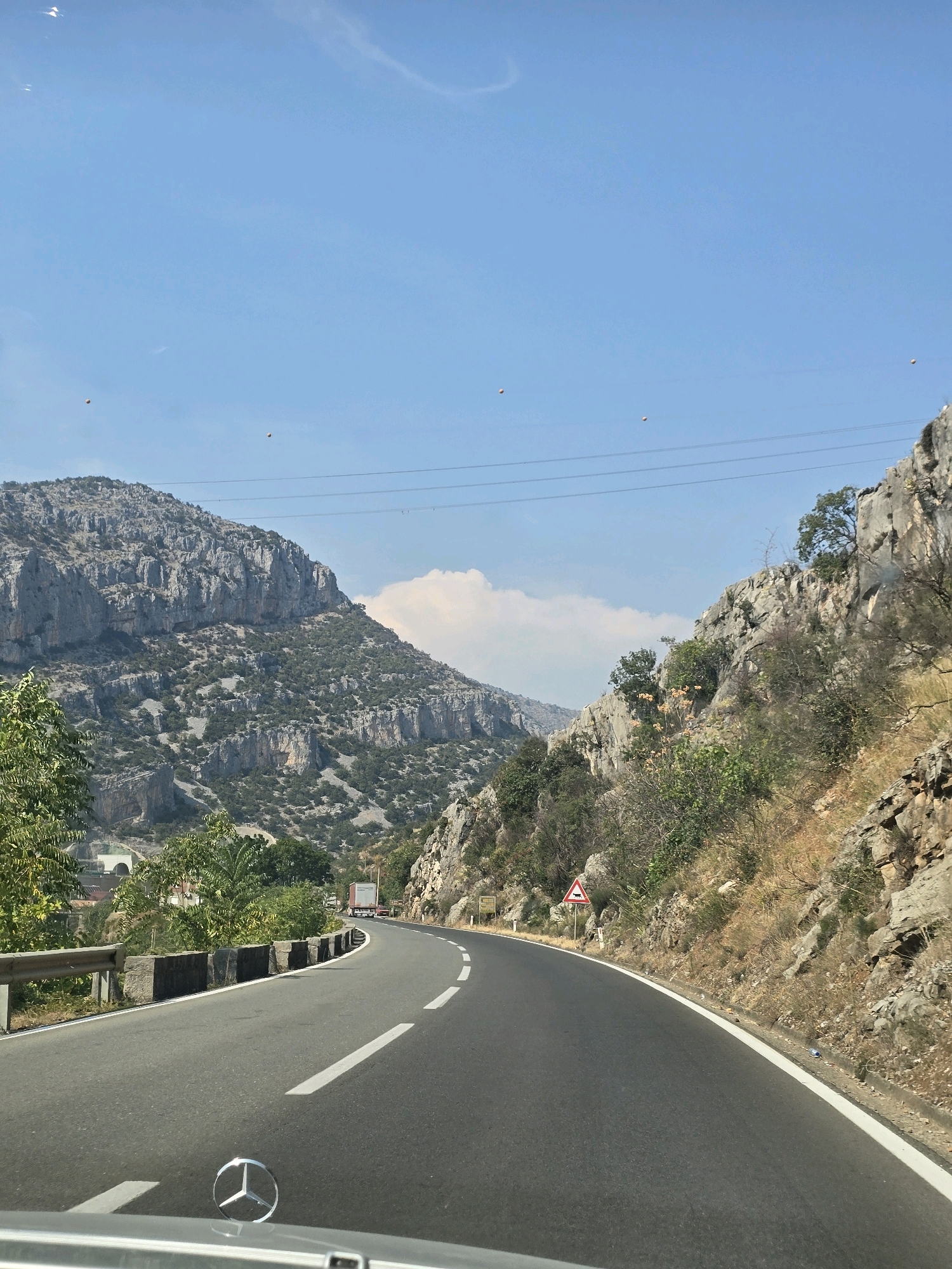 The image shows a winding asphalt road through a mountainous, dry landscape under a clear blue sky. The road curves to the right, with a guardrail on the left side and rocky, vegetated slopes on the right. Several power lines are visible strung across the sky. In the distance, on the left side of the road, there appears to be a tunnel entrance and some greenery. A truck is visible on the road ahead. On the right side of the road, a triangular warning sign with an animal symbol is posted. The foreground shows the hood and Mercedes-Benz emblem of a car, suggesting the photo was taken from a moving vehicle. The overall scene is a scenic mountain pass, likely on a sunny day. This location is not in Podgorica, Montenegro; it appears to be a coastal mountain road.