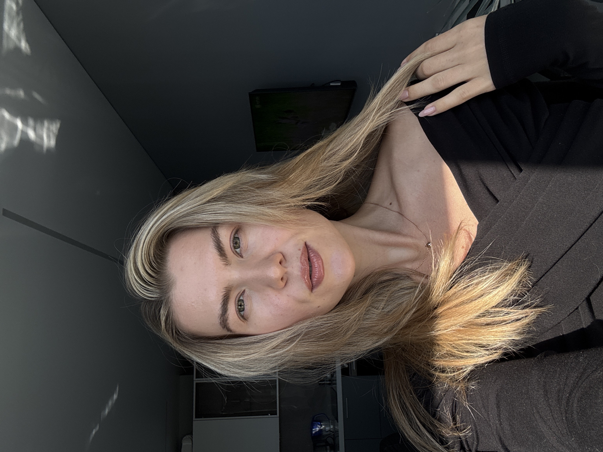 A medium shot of a woman with long, blonde hair. She is wearing a black long-sleeved shirt and a delicate necklace. Her left hand is touching her hair. She appears to be indoors, with a wall visible behind her. The image contains a black TV on the wall. The location in the city of Unknown, Unknown, can not be determined.