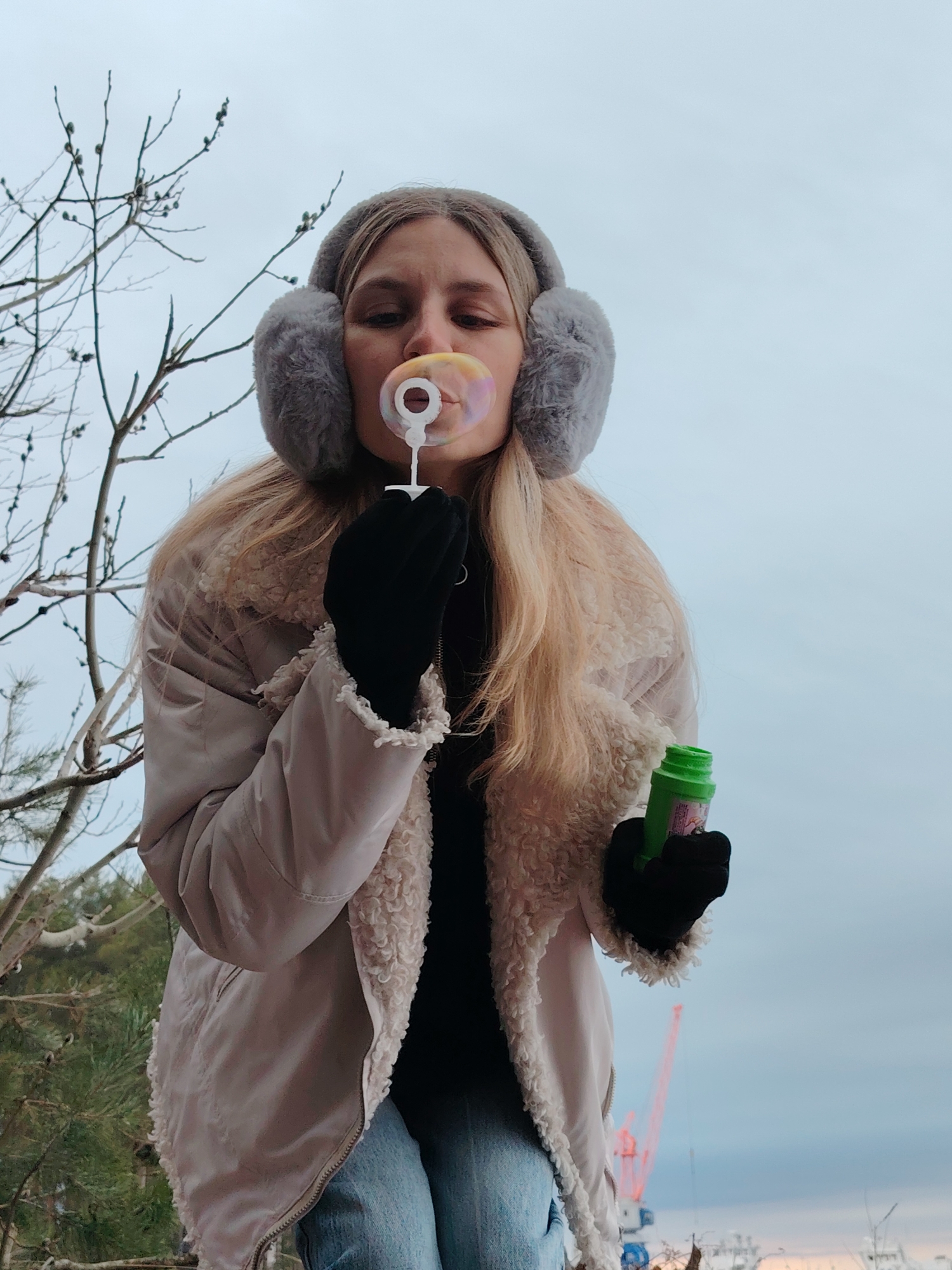 A young woman wearing gray earmuffs, a light beige jacket with a white wool collar, black gloves, and blue jeans is blowing bubbles outdoors. She holds the bubble wand in her left hand and a small green bottle in her right. Bare tree branches are visible to her left, and in the background, there is a cloudy sky, trees, and a red construction crane. The location is Chaikovsky, Russia.