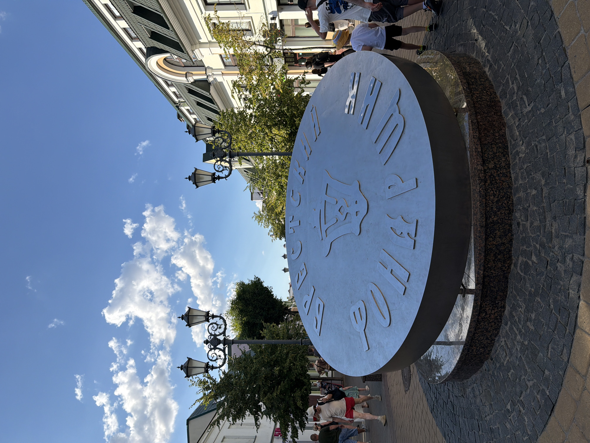 In the city of Unknown, Unknown, a large, circular metal sculpture with Roman numerals and Cyrillic text is positioned in the foreground, angled slightly to the right. The sculpture features a stylized coat of arms in its center. It is set on a circular base made of dark granite with a mosaic pattern of dark and light cobblestones. Behind the sculpture, a street is lined with buildings with green roofs and white facades. Several street lamps with multiple fixtures illuminate the scene. People are walking along the street, some in casual clothing. The sky is blue with scattered white clouds.