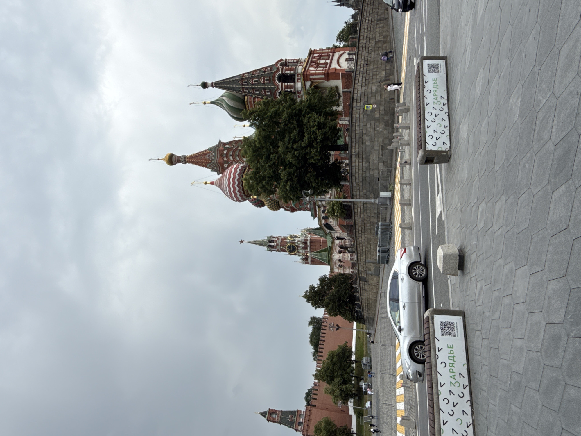 The image depicts a wide, overcast view of Red Square in Moscow, Russia. In the foreground, a grey sedan is driving across a street. To the left of the street, there are two benches with QR codes and Cyrillic text. The background features the iconic St. Basil's Cathedral with its colorful onion domes, the Spasskaya Tower with a clock, and a section of the Kremlin wall. The sky is heavily clouded, suggesting a temperate climate. Several people are visible walking in the midground near the Kremlin wall, and a few cars are parked or driving in the distance. The overall scene captures a moment of everyday activity in a historically significant urban space.