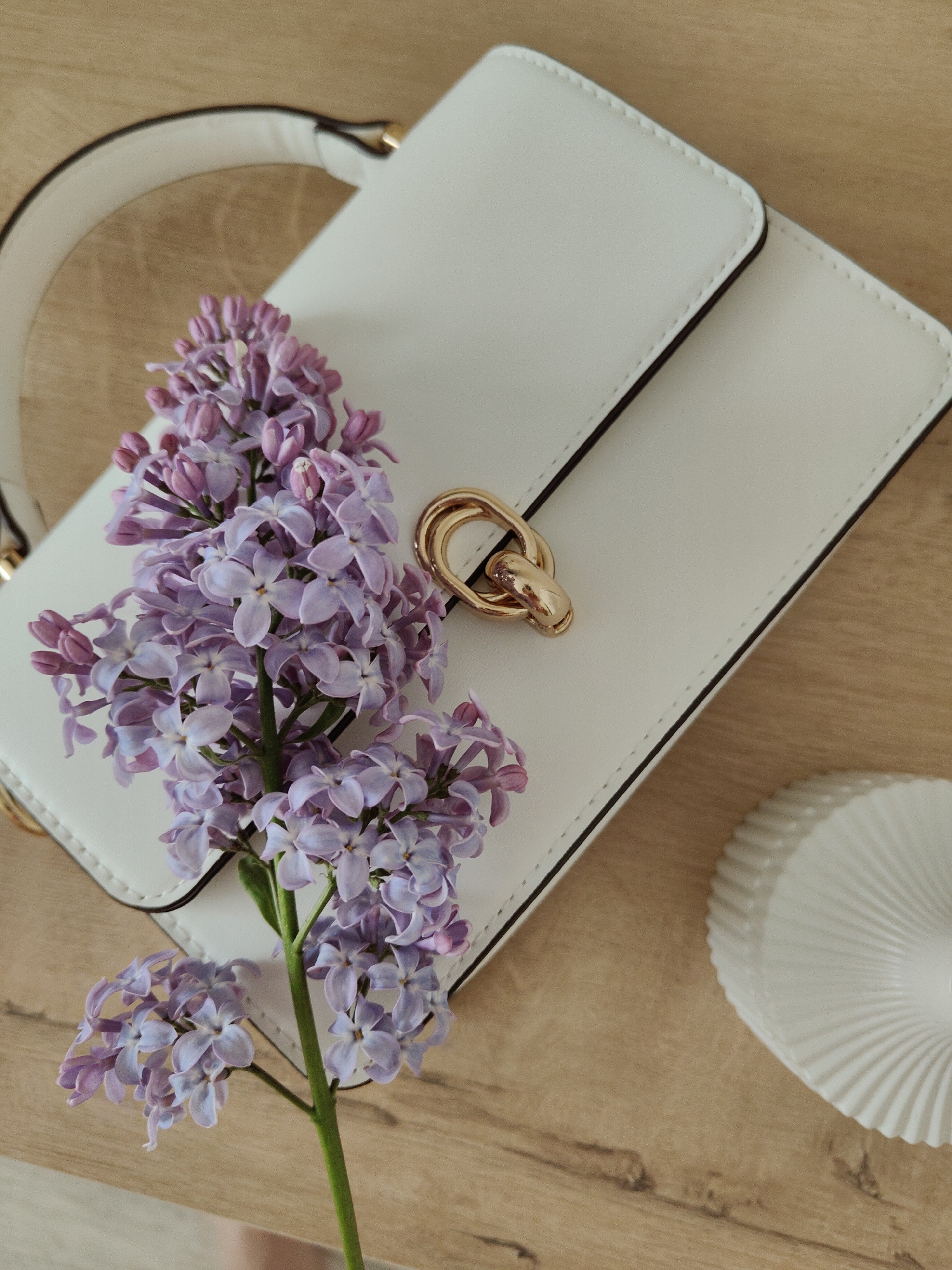 A high-angle, close-up shot captures a white, rectangular handbag on a light-brown wooden surface. The handbag features a gold-toned metal clasp and a white handle. A cluster of lilac flowers rests on the handbag. Beside the handbag is a small, decorative, white object with a ridged, circular design. The location appears to be an interior setting in the city of Unknown, Unknown. There are no people visible in the image. The interaction is a still-life arrangement of the handbag, flowers, and decorative object.