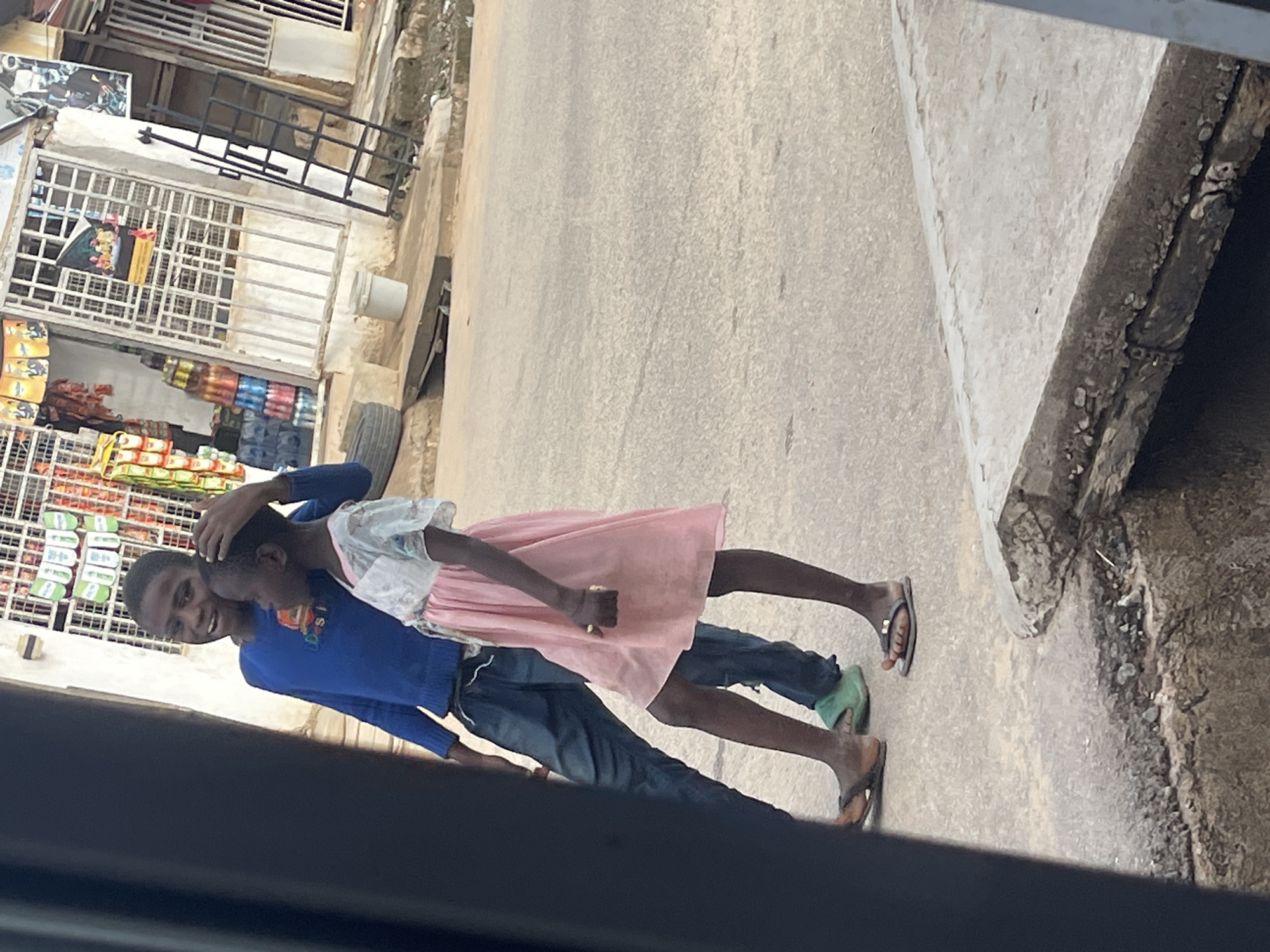 This image depicts a daytime outdoor street scene in a commercial area of Abuja, Nigeria. The foreground features two dark-skinned children positioned on a light gray paved surface.
On the left, an older male child, wearing a long-sleeved blue top with partial text "D...s I" visible on the chest, and dark trousers, stands with his left hand resting on the head of a younger female child to his right. He is wearing brown flip-flops and displays a smile directed towards the viewer.
The younger female child, dressed in a light-colored sheer top over a pink knee-length dress, holds a small yellow object in her right hand. She is wearing dark flip-flops with a metallic embellishment and is oriented in profile, looking towards her left.
The children stand near the edge of a concrete drainage ditch that runs along the side of the paved surface. The ditch is partially covered by a concrete slab, with exposed sections and some debris visible, particularly towards the bottom right.
In the background, behind the children, are white-painted buildings with prominent metal security grilles on their windows and doorways. The building to the left features racks with hanging packaged goods, banners, and stacks of multicolored bottled drinks. A white plastic bucket and a dark vehicle tire are visible near its base. To the right, another building has a black metal, ladder-like structure leaning against its facade, with additional posters visible. The ground in the far right background appears rough and unpaved.