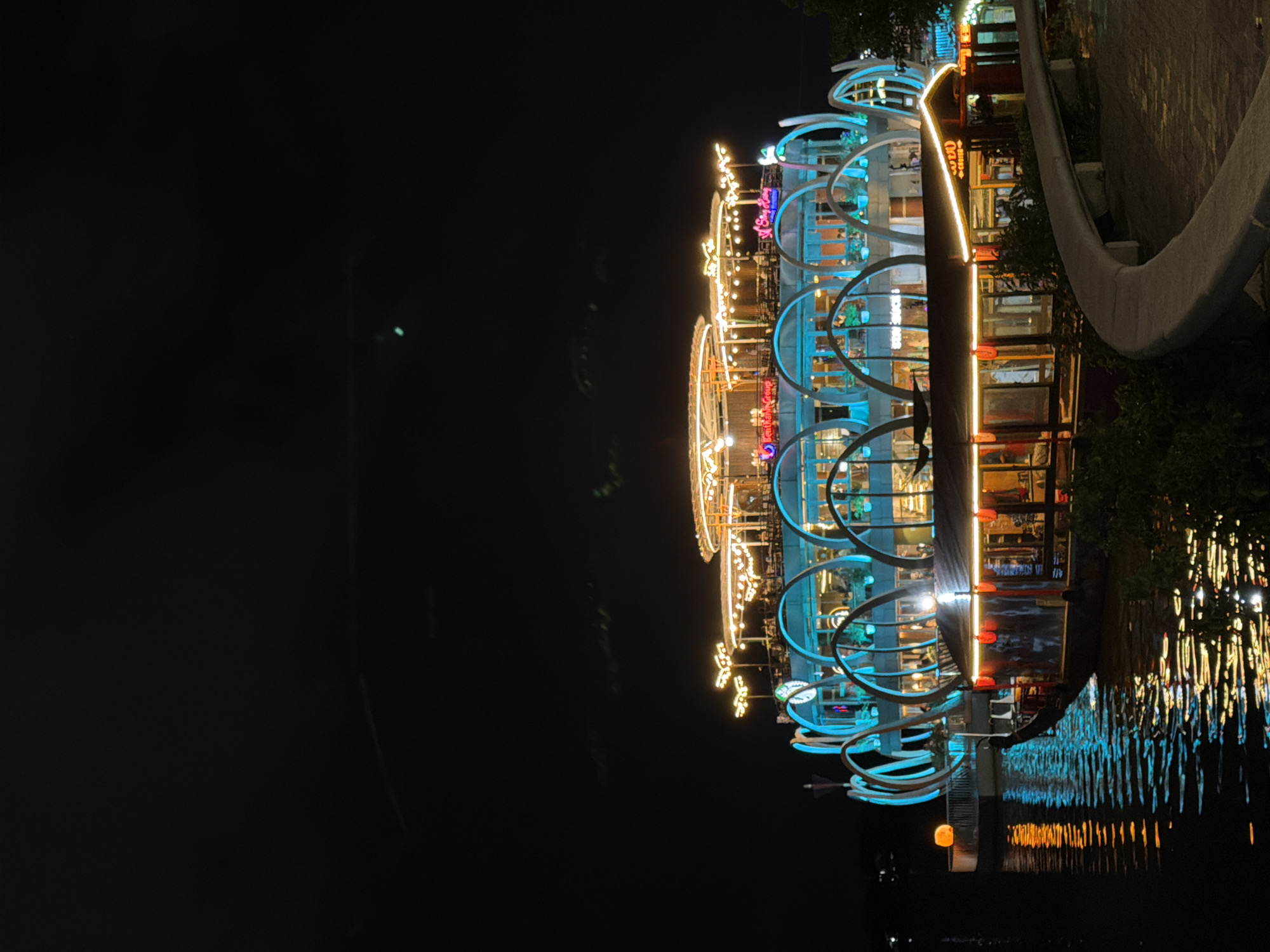 At night in Phú Xuân Ward, Vietnam, a brightly lit, multi-level boat restaurant is docked on a body of water. The boat has a unique architectural design featuring several tiered levels adorned with a multitude of warm-toned string lights. The main structure of the boat is illuminated with vibrant blue arched accents, creating a striking contrast against the dark sky. The lower deck of the boat is visible, featuring windows with interior lighting and traditional red lanterns hanging from the eaves. The water surrounding the boat reflects the lights from the boat and the sky, creating a shimmering effect. A curved concrete walkway is visible in the foreground, suggesting a waterfront promenade. The sky above is dark with some faint cloud formations.
