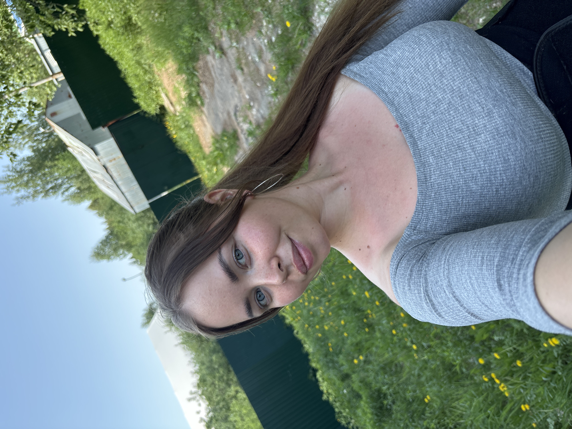 A selfie of a woman with brown hair and blue eyes. She is wearing a gray long-sleeved shirt and a black jacket or backpack. The background shows a grassy area with yellow flowers and a shed or building constructed of corrugated metal. The location appears to be in Русский Вожой, Russia, based on the prompt.