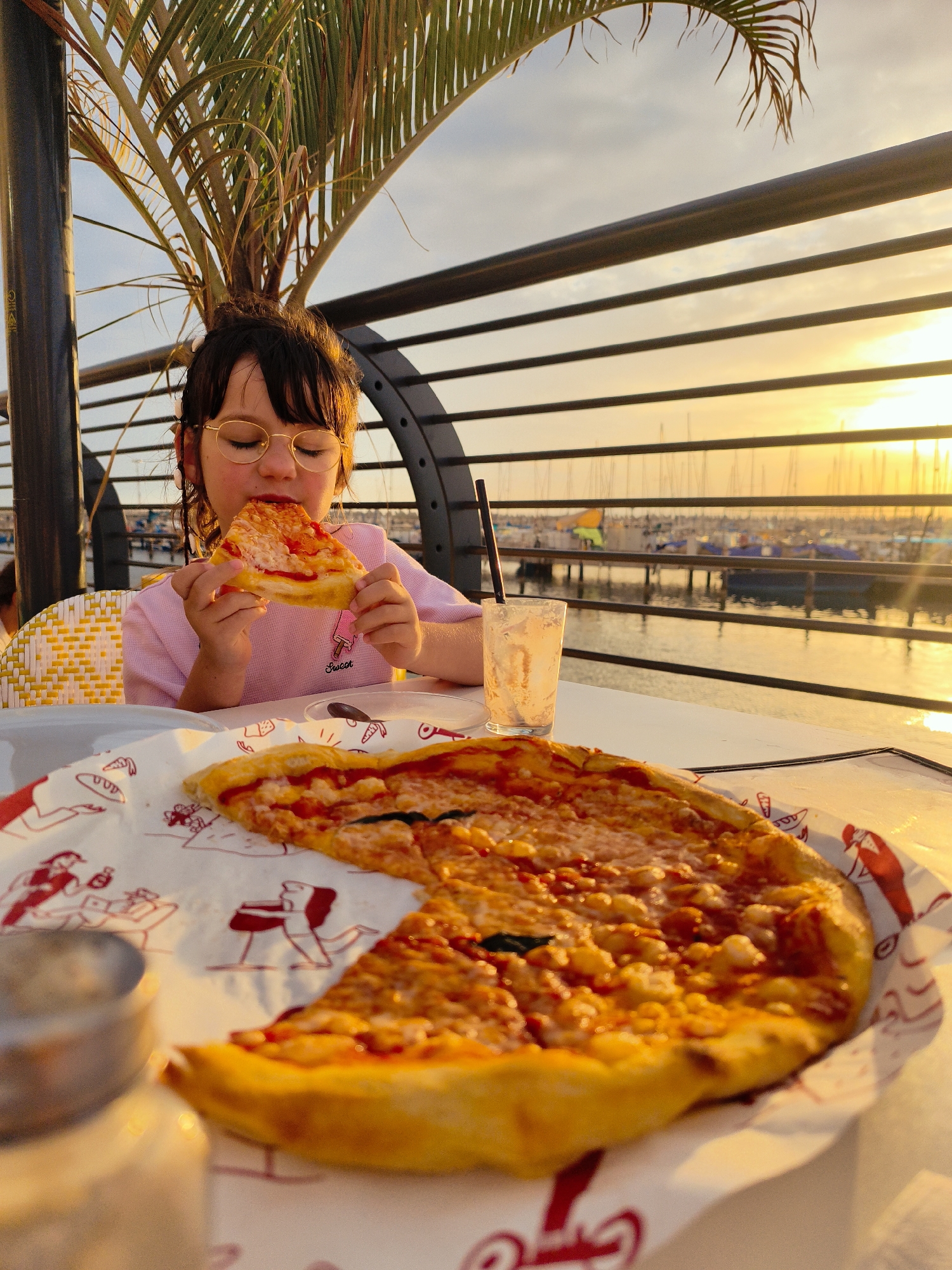 A young child with dark hair and round, gold-rimmed glasses is the central subject of this vibrant outdoor image. The child, wearing a pink t-shirt with the word "Sweet" visible on it, is actively eating a slice of cheese pizza, holding it up to their mouth with both hands. Their expression suggests focused enjoyment of the meal.
In the foreground, a large, partially eaten cheese pizza rests on a white paper liner adorned with red, cartoon-style illustrations of people. Next to the pizza, on a white table, is a half-empty glass with a black straw. Part of a metallic object is visible on the far left of the table.
The setting is an outdoor dining area, likely a restaurant or cafe situated by a marina or port. In the background, numerous boats are docked in calm water, stretching into the distance. A metal railing with horizontal bars separates the dining area from the water. Above the railing, the sky glows with warm, golden hues of sunset or golden hour, casting a bright, serene ambiance over the scene. A vibrant green palm tree frond is visible in the upper left, adding a tropical element. The warm light creates strong reflections on the water and highlights the details of the pizza and the child's face. The overall impression is one of a peaceful and enjoyable evening meal by the water, likely in Ashkelon, Israel.