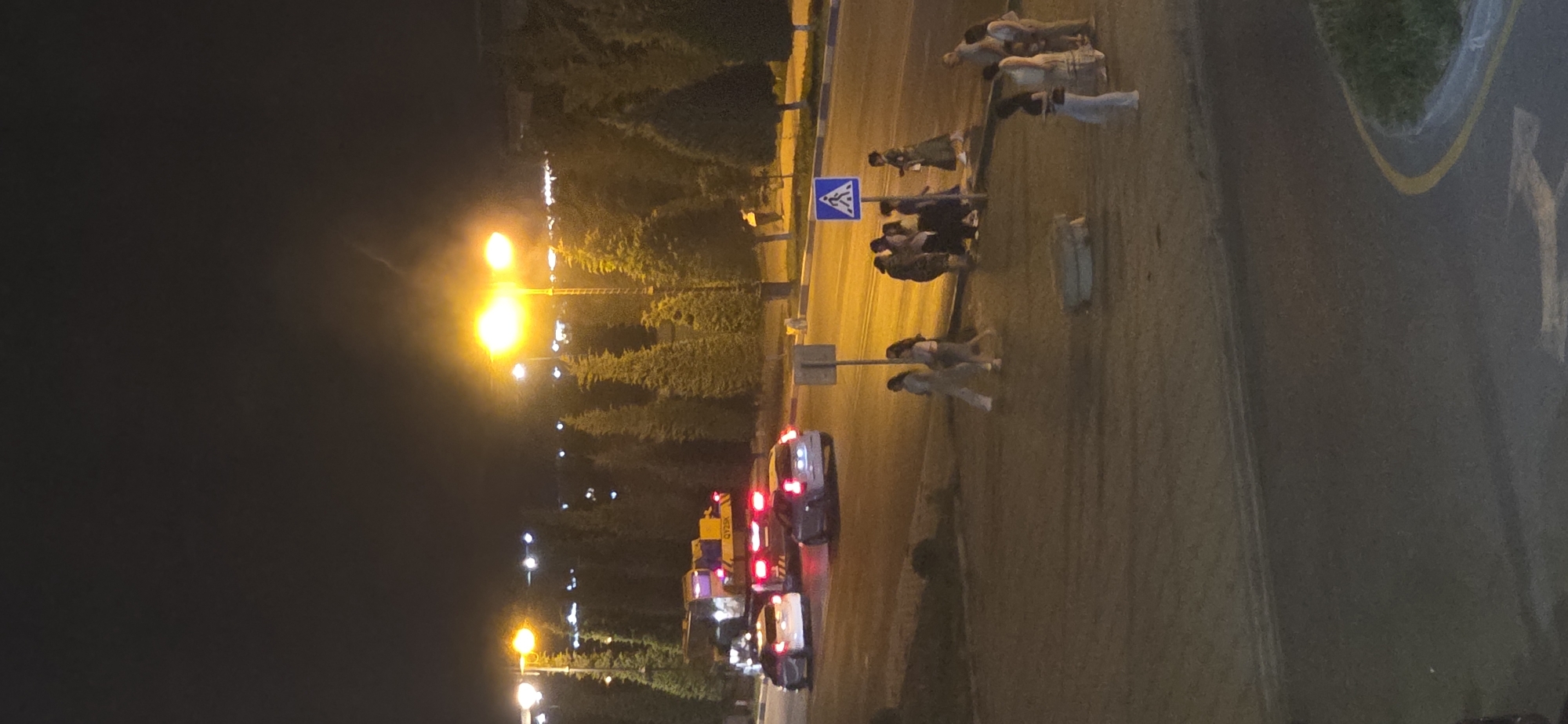 This is a night-time, vertical, medium-distance shot of a street scene in Sumgayit, Azerbaijan. The sky is dark, with a bright, artificially lit area in the upper right quadrant, possibly from streetlights or a building. The foreground is a paved area, with a dark, asphalt road curving to the right. On the road, there are several vehicles, including a white car, a dark grey car, and a yellow and blue truck or large vehicle. In the background, there are rows of evergreen trees.
Further into the scene, on the right side of the road, there is a pedestrian crossing with a blue and white "pedestrian crossing" sign. A group of people, appearing to be mostly women, are walking across the street or standing near the crossing. Some are dressed in light-colored clothing, while others are in darker attire. To the far right, there are more people standing. There are several streetlights visible, casting a bright yellow glow, illuminating parts of the scene. The overall impression is a busy urban environment at night.
