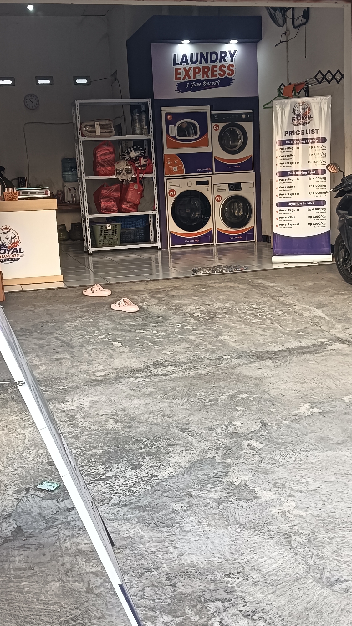 The image shows the exterior of a "Laundry Express" business located in Sragen, Indonesia. A prominent sign with the business name is visible, along with several washing machines. To the left of the laundry machines, there is a shelf unit filled with laundry bags and water bottles. In front of the laundry service, a price list is displayed. The floor is concrete and appears to be an outdoor area, possibly a sidewalk or storefront entrance. Two pairs of pink flip-flops are lying on the ground in front of the laundry area. The overall environment suggests a commercial establishment in a public space.
