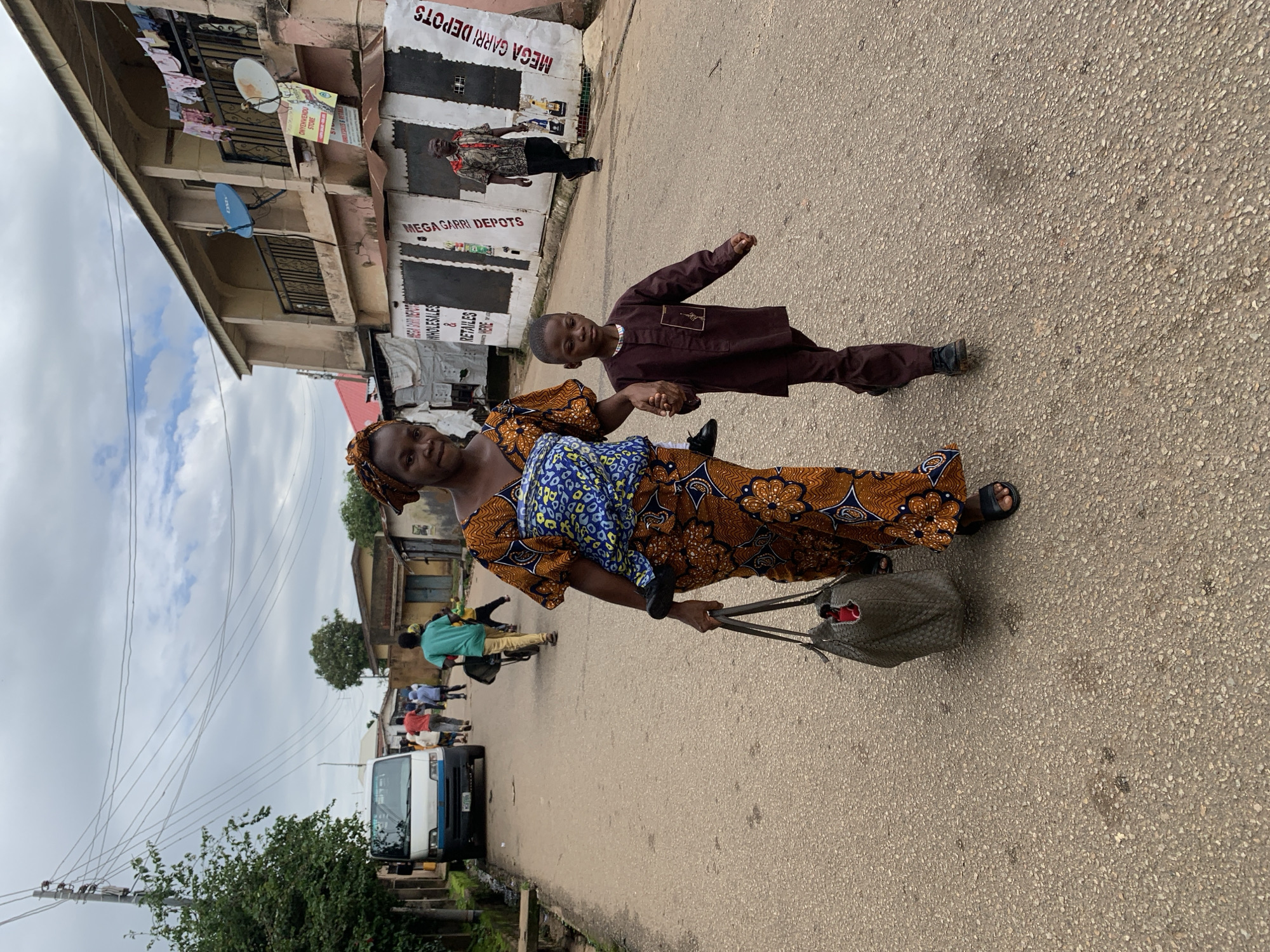 In Jos, Nigeria, a woman and a young boy walk on a paved road under an overcast sky. The woman, carrying a baby on her back in a blue and yellow patterned fabric sling, wears a vibrant orange and brown patterned dress and a matching headwrap. She holds a dark woven tote bag in her left hand and the boy's right hand with her right hand. The boy, dressed in a brown tunic and trousers, walks beside her. In the background, several other people are visible walking in the same direction, along with a white and blue van and a few buildings. One building, to the right of the frame, displays signs for "MEGA GARRI DEPOTS" and "CHYENWENOU STORE." Clothes hang from a balcony on the upper floor of this building. The road surface is rough asphalt, with scattered debris.