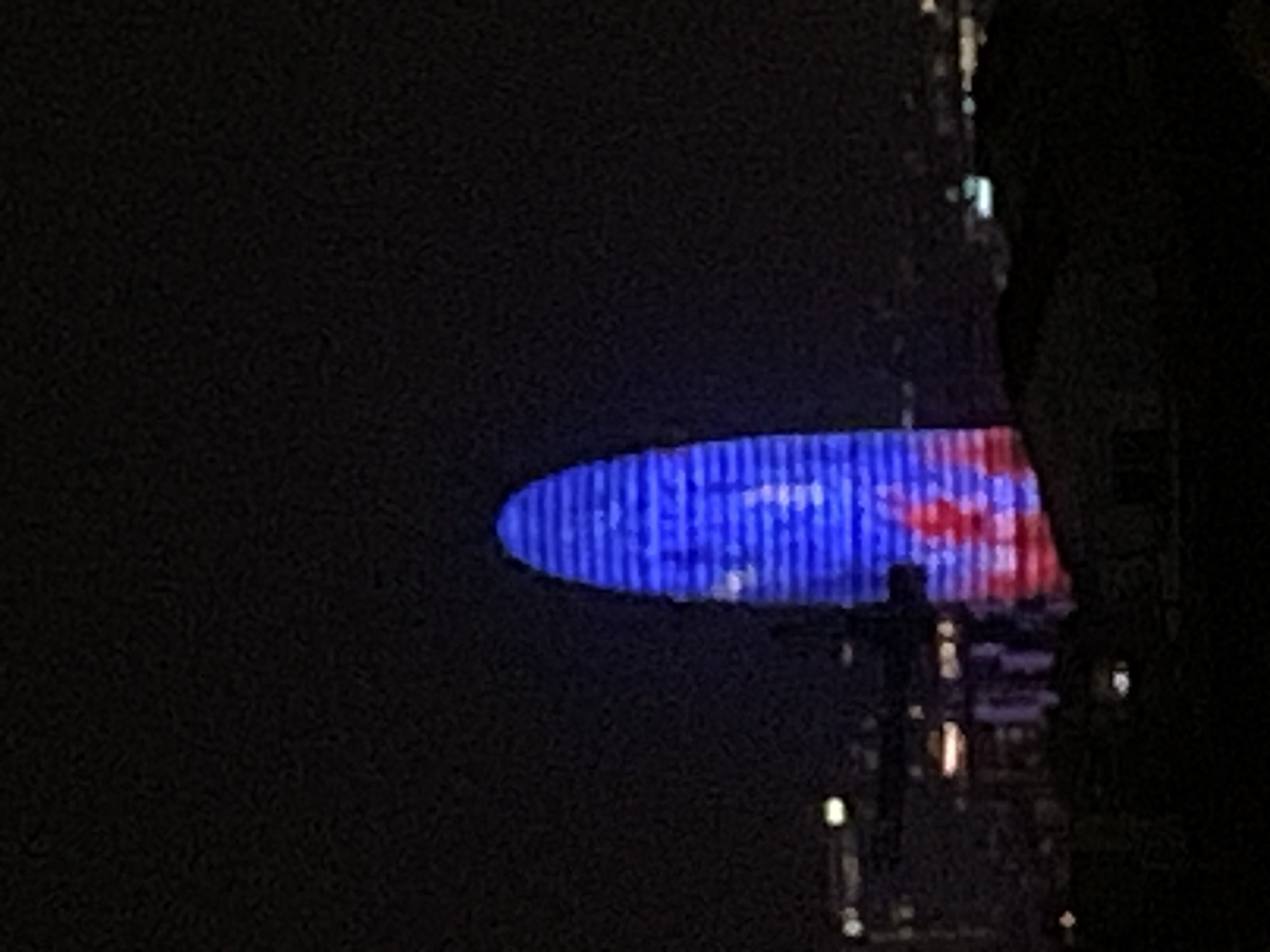 The image shows the Agbar Tower in Barcelona at night. The tower is illuminated with blue and red lights. Other buildings are visible to the right of the tower. The image appears to have been taken from a distance.