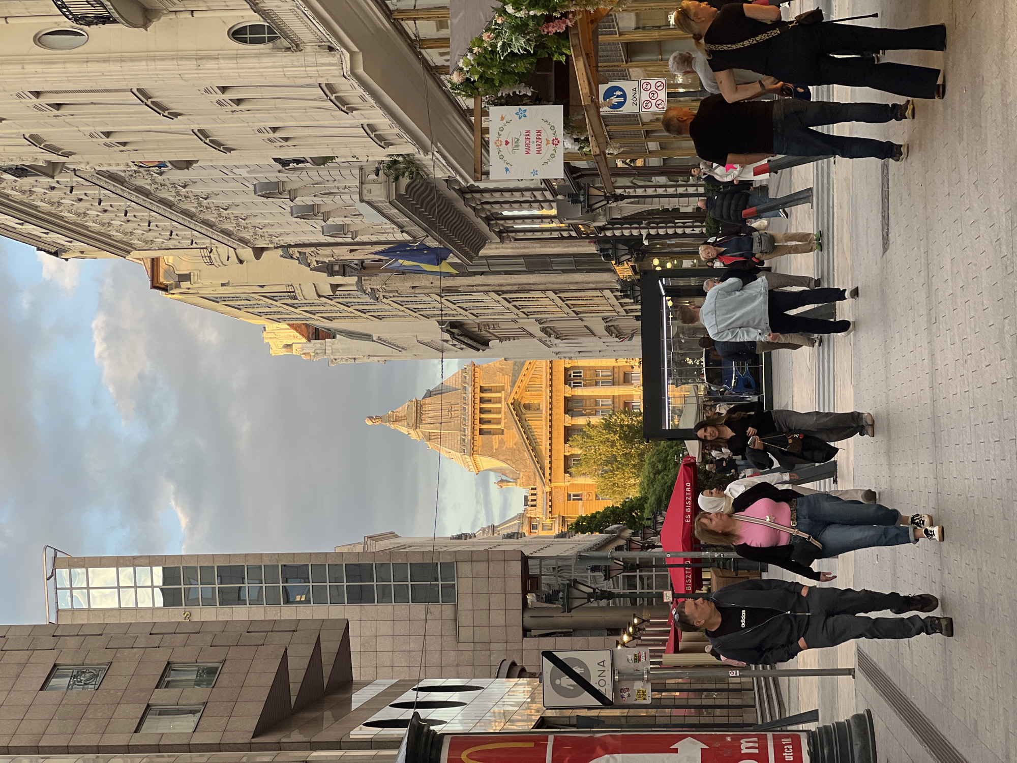 This image depicts a street scene in Budapest, Hungary. The view is of a pedestrianized street lined with buildings of various heights and architectural styles. People are walking on the street, including individuals and groups. Some are casually dressed, while others are wearing more formal attire. There is a McDonald's sign at the bottom of the image, along with a sign indicating a pedestrian zone, “ZÓNA.” There is also another sign for "TAMS MARCIPÁN MARZIPAN." In the distance, a large, ornate building with a prominent dome can be seen. The sky is overcast with clouds.