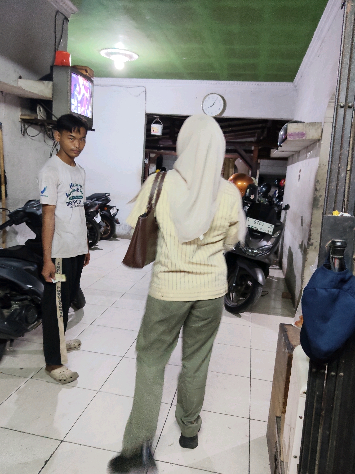 The image depicts an indoor scene, likely a garage or a covered area for motorcycles, in Purwakarta, Indonesia. A young man in a white t-shirt and black pants stands on the left, holding a wad of cash. He is facing the camera with a neutral expression. To his right, a person wearing a beige striped long-sleeved shirt and a light-colored hijab is walking away from the camera, carrying a brown bag over their shoulder. The floor is tiled with a white and grey grid pattern. Several motorcycles are parked in the background. The environment appears somewhat cluttered, with a television set mounted on the wall to the left and a clock visible on the upper wall. The lighting suggests it might be indoors or a covered outdoor space.
