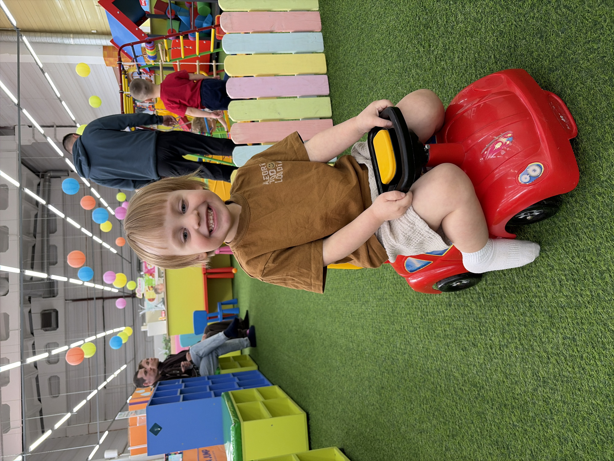 A child is sitting in a red toy car in an indoor playground, likely in Chaikovsky, Russia. The child is fair-skinned with blond hair, wearing a brown t-shirt and light-colored shorts with white socks. The child is smiling at the camera. The car is red with black wheels and a black and yellow steering wheel. The floor is covered in green artificial turf. In the background, there are adults, other children, colorful structures, and play equipment, including a colorful wooden fence and shelving. The ceiling has a white, grid-like structure with colorful hanging paper lanterns.