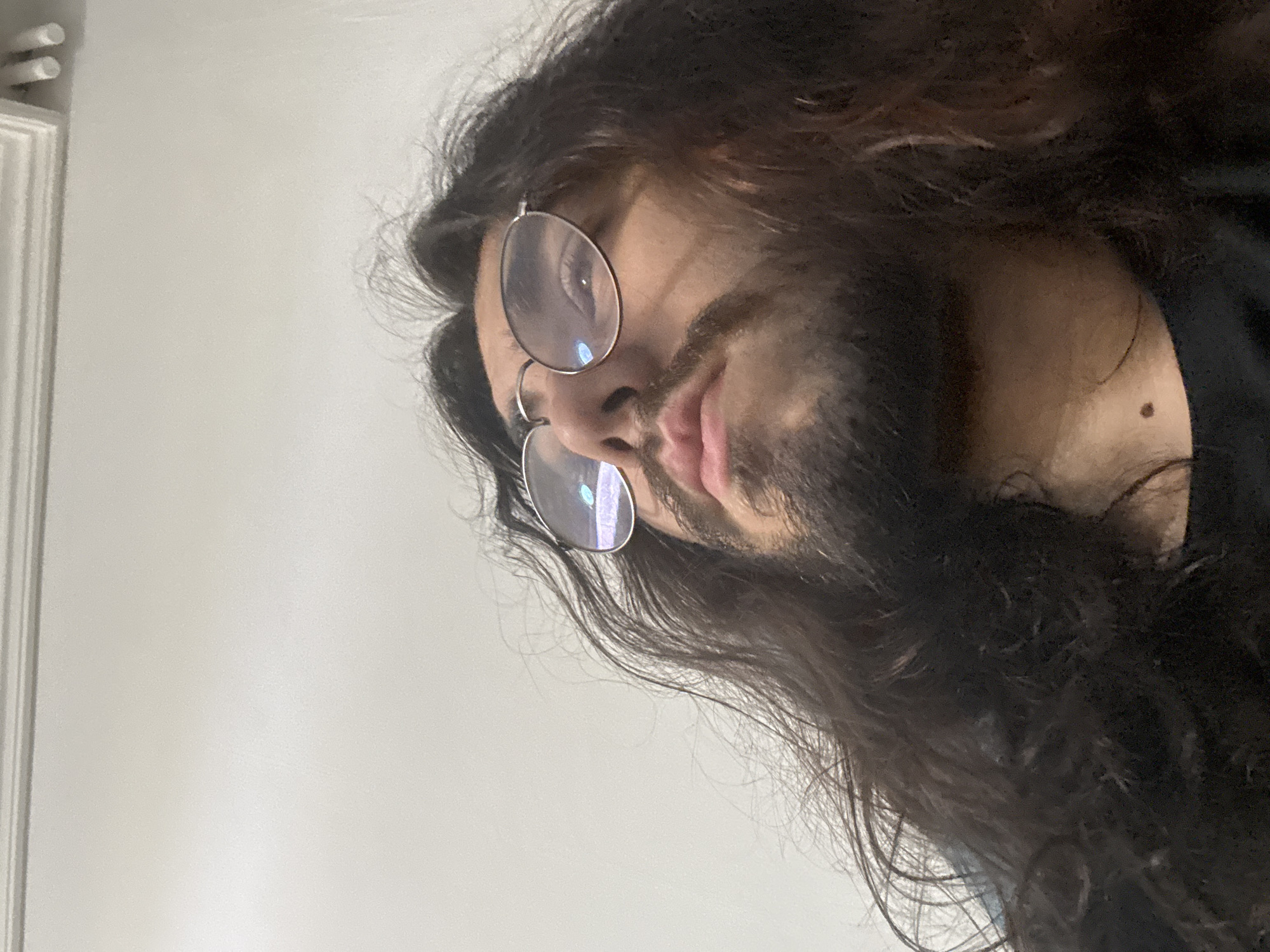 A close-up, low-angle, indoor shot shows a person with long, dark, wavy hair and a beard. They are wearing round, wire-rimmed glasses. The person's gaze is directed upwards and to the left. The background consists of a plain, light-colored wall with molding near the top. The lighting appears to be artificial. No specific location in the city of Unknown, Unknown can be determined from the image.