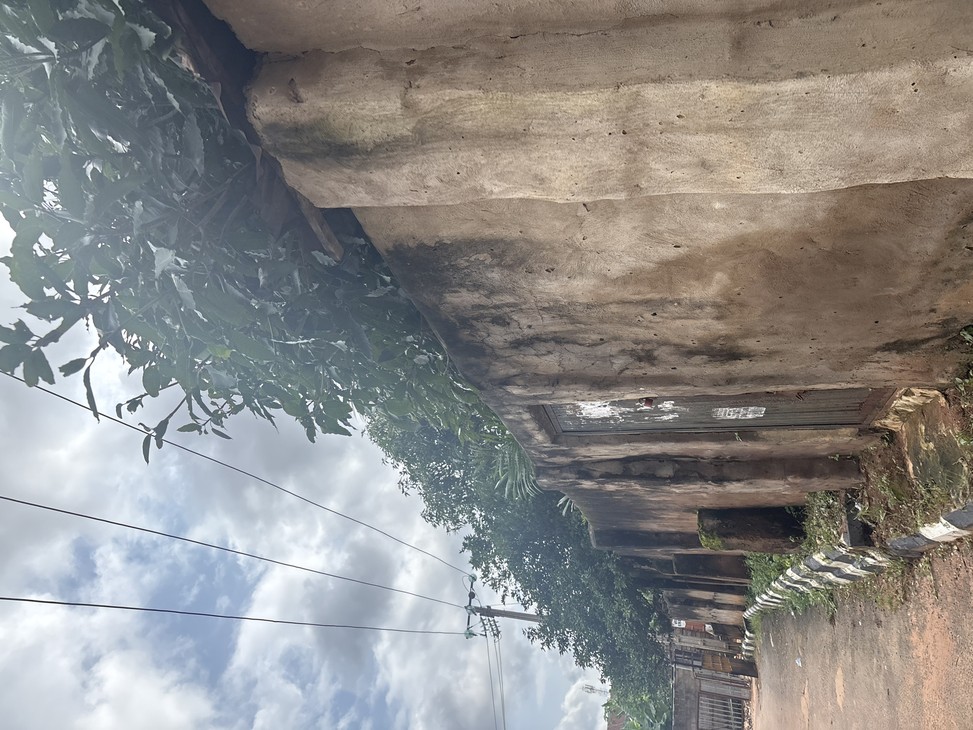 This image captures a section of a street in Nkpor, Nigeria. The foreground is dominated by a textured, weathered concrete wall that extends diagonally across the frame, with a weathered wooden door set into it. The wall appears aged, showing signs of staining and erosion. Above the wall, lush green foliage overhangs, with a wooden beam visible at the top. In the mid-ground, a paved road curves to the left, bordered by a kerb with alternating black and white stripes, suggesting a demarcation. Greenery and some rubble are visible along the kerb. Further down the road, a metal gate and fence can be seen. Electrical wires stretch across the sky, supported by a utility pole in the background, which has insulators and crossbars. The sky is filled with a mix of white and grey clouds. The overall impression is one of an urban environment with signs of wear and tear, common in many developing areas.