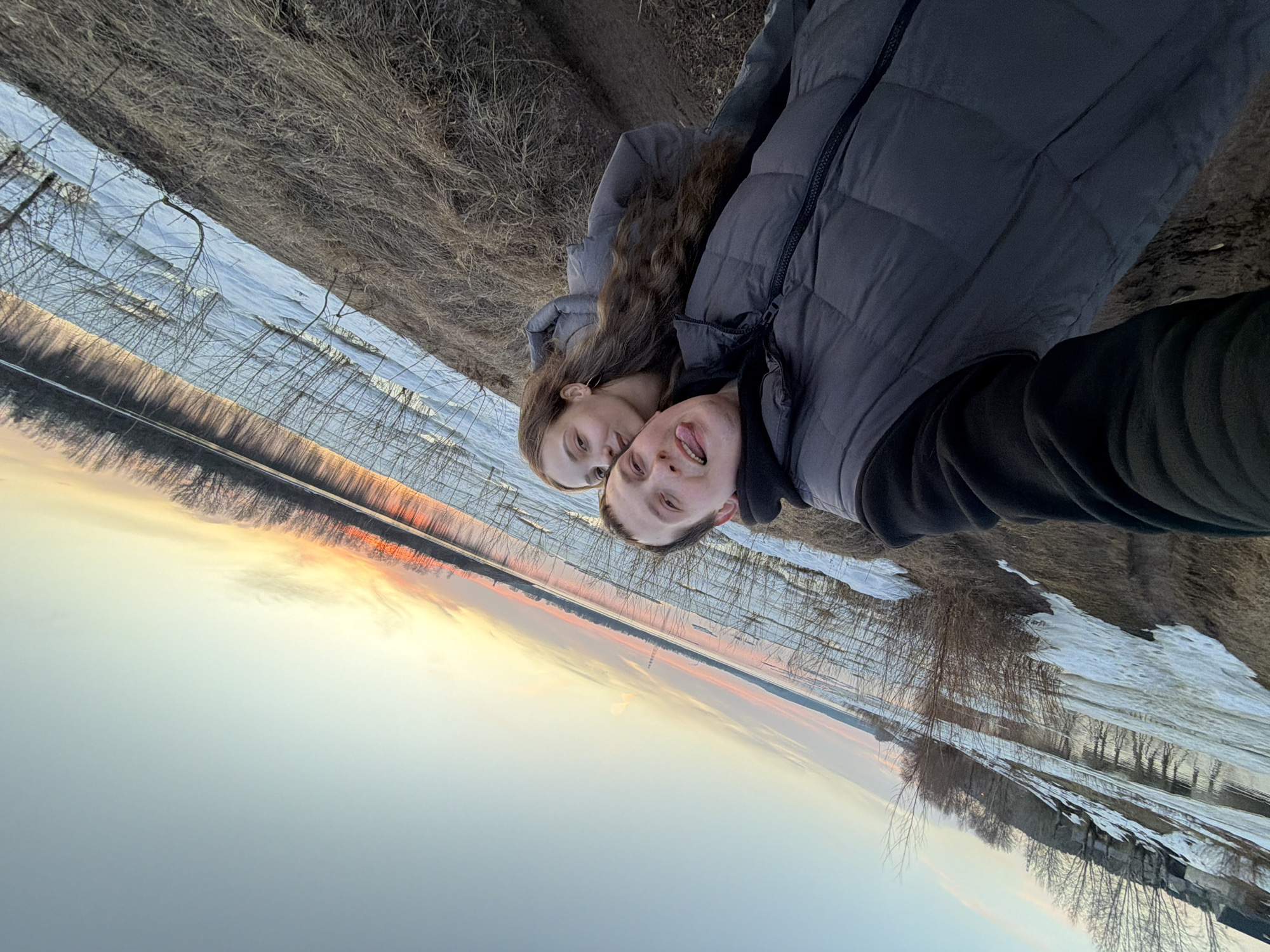 A selfie shows a young couple outdoors. The man, in a dark gray puffer jacket and black sweatshirt, sticks his tongue out. The woman, with long brown hair, wears a gray puffer jacket. They stand in front of a landscape, likely during the sunset. There is water, possibly a river, reflecting the colors of the setting sun. Bare trees are visible on the opposite bank. Snow is on the ground in patches. Based on the landscape, this image was taken near Chaikovsky, Russia. The location might be near a river or body of water with snow on the ground and bare trees.