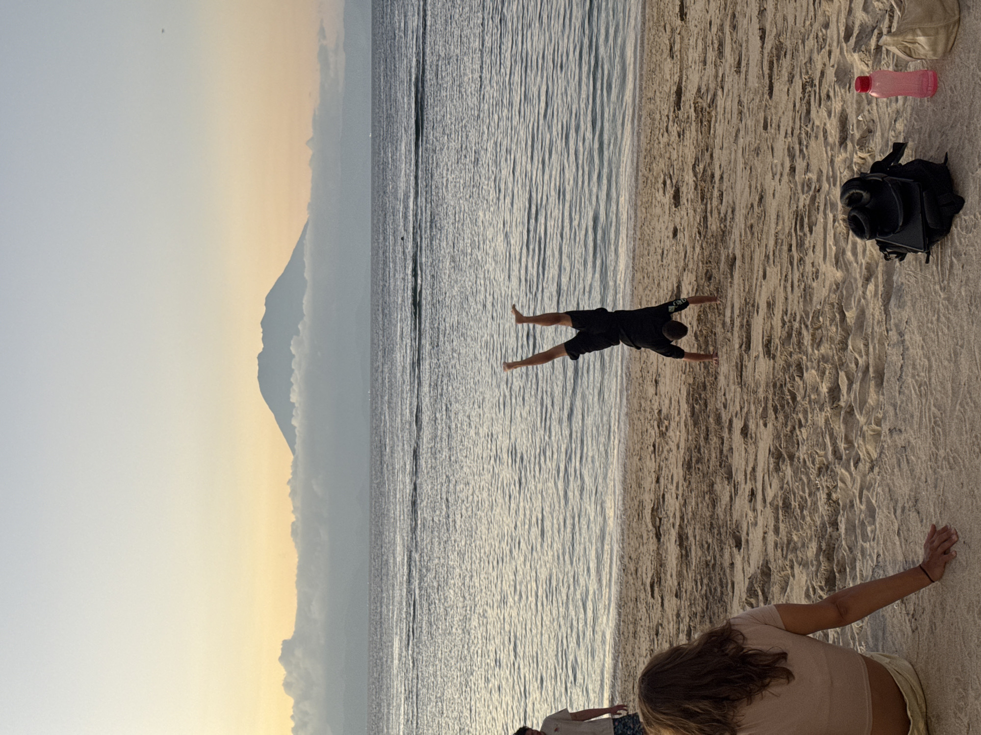 This image captures a vibrant beach scene in Nusa Penida, Indonesia, during what appears to be late afternoon or early evening, bathed in warm, soft light.
In the foreground, to the right, a person with long brown hair, wearing a light-colored top and shorts, is seen from behind, leaning or sitting on the sandy beach. Their right arm is extended, resting on the sand. Further to the right, on the sand, are several personal items: a black backpack, a bright pink water bottle, and a light-colored fabric bag. The sand itself is textured with ripples and footprints.
The midground features the tranquil ocean, characterized by gentle ripples reflecting the soft light. A male figure, dressed in dark shorts and a dark shirt, is captured in a dynamic pose resembling a handstand or a dive at the water's edge. His body forms a 'V' shape, with arms extended forward and legs spread slightly apart, creating an energetic focal point against the water.
In the background, a large, majestic mountain or volcano rises prominently on the distant horizon, partially obscured by a soft, atmospheric haze. The sky transitions from a pale blue at the top to a warmer, yellowish-orange gradient closer to the horizon and the mountain, indicative of the golden hour. Given the location context of Nusa Penida, this mountain is most likely Mount Agung on Bali.
The overall scene conveys a sense of leisure, natural beauty, and playful activity by the sea. There is no visible text in the image.