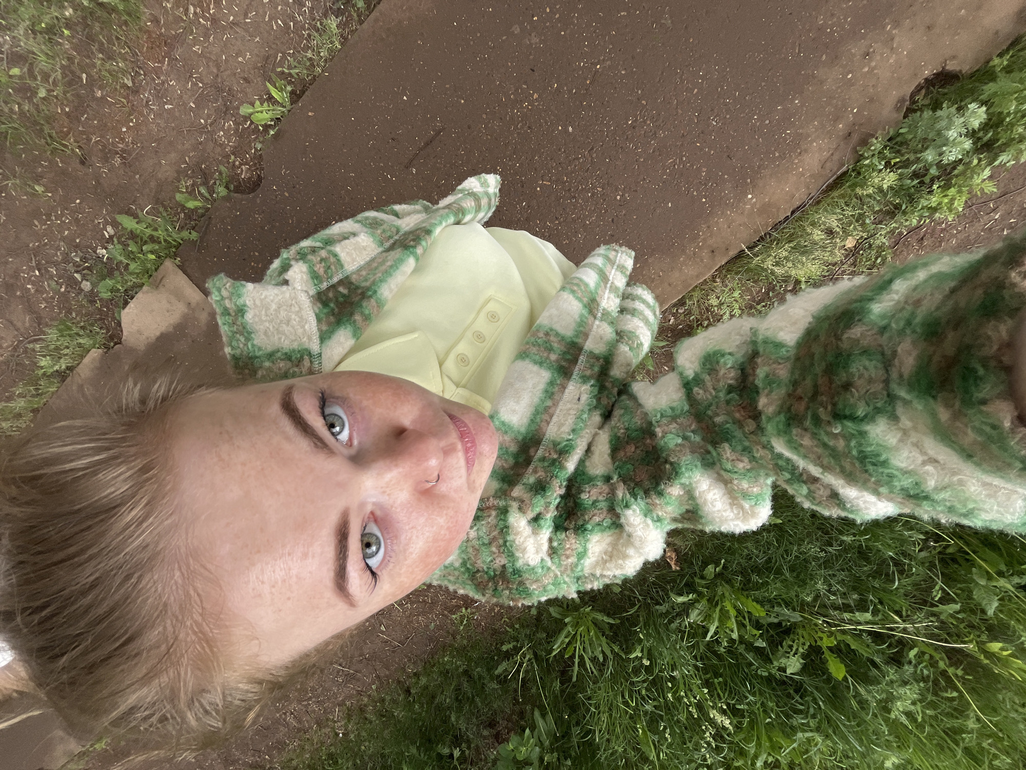 A high-angle, top-down selfie captures a young adult outdoors, looking directly up at the camera. The subject, likely female, has light blonde or brown hair and striking blue-green eyes. Her face shows light freckles across the nose and cheeks, and a small silver hoop nose ring is visible on her right nostril. She is wearing a light yellow collared shirt with three visible buttons down the front, layered under a chunky, fuzzy jacket. This jacket features a distinct green and off-white plaid or checked pattern.
The background depicts an outdoor natural setting with varying textures. Directly below and to the left of the subject, there is dark brown soil with scattered small green plants and weeds. A diagonal strip of darker, granular material, resembling an asphalt path or concrete edge, cuts through the lower and middle portions of the frame, separating the dirt from a denser area of wild green grass and foliage to the right. The lighting appears soft and even, suggesting it is daytime, possibly under overcast skies, as there are no harsh shadows. The person's expression is neutral and direct. The image suggests a casual moment captured in a garden, park, or natural area in Chaikovsky, Russia. No visible text is present in the image.