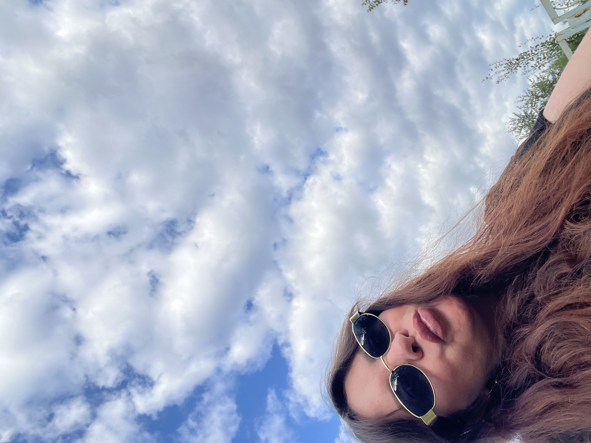 A low-angle, close-up selfie of a woman with long, brown hair and sunglasses is captured against a cloudy sky. The woman is wearing dark sunglasses with gold accents and her lips are pursed. Her hair is flowing loosely around her shoulders and down her back. The background consists of a blue sky with scattered white clouds, suggesting an outdoor setting. There are no visible landmarks or indicators that place the image specifically in Noviy, Russia.