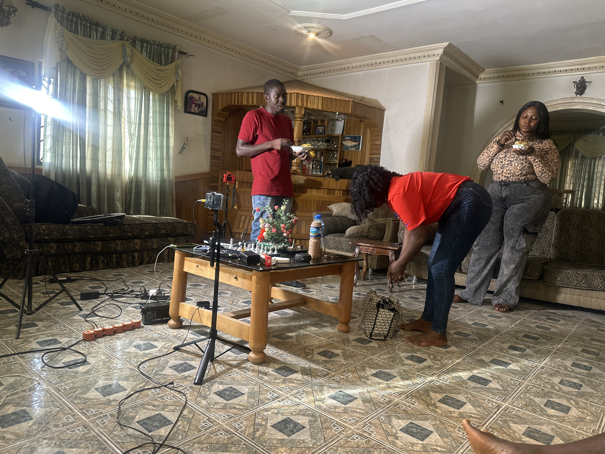 The image depicts an interior scene, likely a living room, in Jos, Nigeria. Three individuals are present: A man in a red shirt stands near a built-in display case, holding a plate. Another individual, wearing a red shirt and jeans, is bending over near a coffee table. A woman in a leopard-print top and gray pants is standing and eating from a bowl. The floor is tiled with a geometric pattern. A coffee table in the center is covered with objects, including a chess set, flowers, and bottles. Camera equipment and power strips are scattered on the floor near the table. A light stand is positioned near a window covered with curtains. Upholstered sofas are placed around the room. The room is lit by both natural light from the window and artificial lighting.