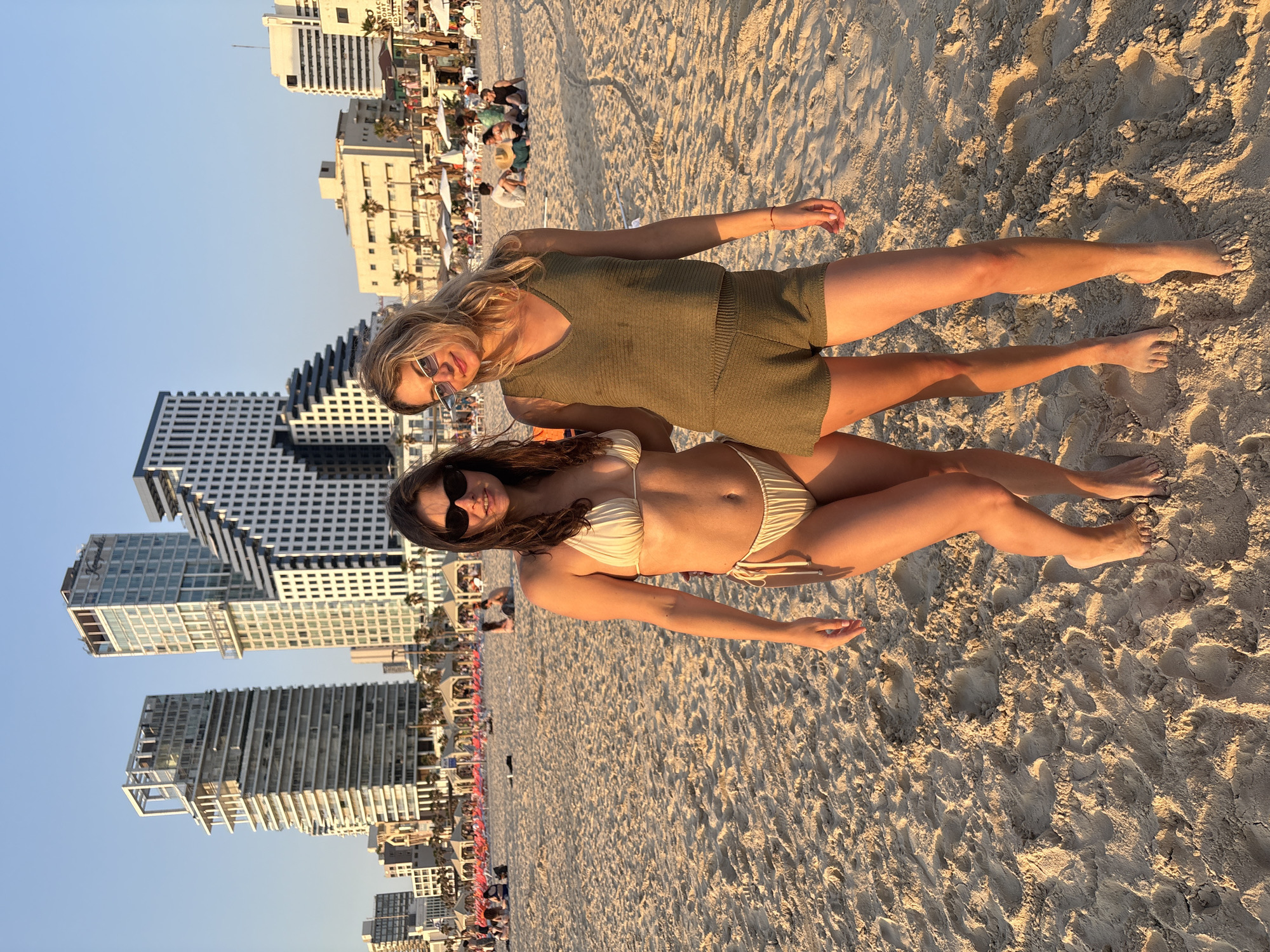 Two women stand on a sandy beach in Tel Aviv, Israel, bathed in warm, golden light during the late afternoon or early evening. The woman on the left, with dark hair and sunglasses, wears an off-white two-piece bikini and smiles at the camera. The woman on the right, with lighter hair and rectangular sunglasses, wears an olive green knit V-neck tank top and matching shorts, also smiling at the camera. Both are barefoot, standing on textured sand with visible footprints. In the midground, the beach is populated with numerous people relaxing on lounge chairs, many covered in red or pink fabric, and under umbrellas. Several beachfront structures, likely cafes or restaurants, are also visible. The background showcases the distinct Tel Aviv skyline, featuring prominent buildings such as the David Kempinski Tel Aviv hotel, the Royal Beach Tel Aviv hotel, and the Andromeda residence, confirming the location. The sky is clear and light blue, indicating fair weather.