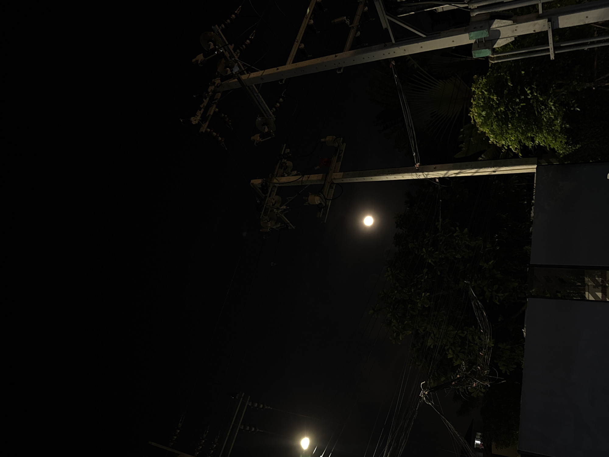The image presents a nighttime scene dominated by urban utility infrastructure. The sky is dark, with a prominent, circular, bright light source (likely the moon or a strong street light) situated in the upper-middle right portion of the frame. A second, fainter light source glows in the bottom-left corner.
Several utility poles and a dense network of overhead power lines, electrical cables, insulators, and transformers diagonally cross the image from the top-right to the mid-left. These structures are largely silhouetted against the dark sky, though some components reflect light.
Green foliage from trees is visible along the right side of the image and peeks from behind the utility structures. Distinct palm-like fronds are noticeable within the greenery. A dark, rectangular structure, possibly a section of a building or a large sign, occupies the far right edge.
The scene captures the complex and intricate web of wires and poles common in many urban landscapes, particularly at night. No people or specific activities are depicted. The time of day is clearly night, under clear weather conditions. No visible text is present. The setting strongly suggests an urban or semi-urban environment, aligning with the context of Bangkok, Thailand.