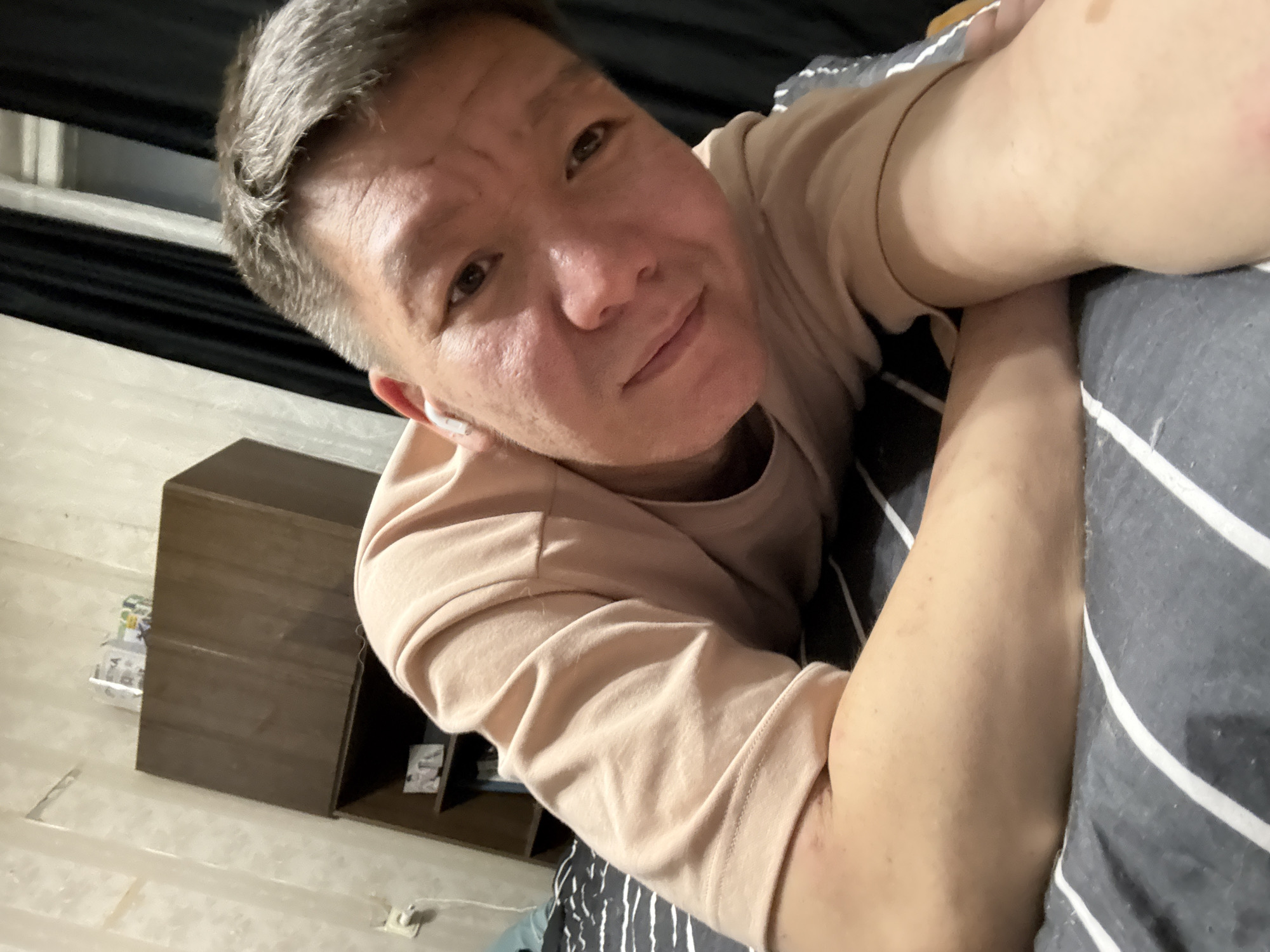 A young Asian man with short, graying hair and fair skin is lying on a bed, taking a selfie. He is wearing a light peach t-shirt and white wireless earbuds. His left arm is bent and resting on the bed, with his hand near his face. The background shows a dark curtain and a wooden shelf unit with some items on top. The bed has striped gray and white bedding. The overall impression is that of a casual, indoor setting, possibly a bedroom in an apartment in Khimki, Russia.