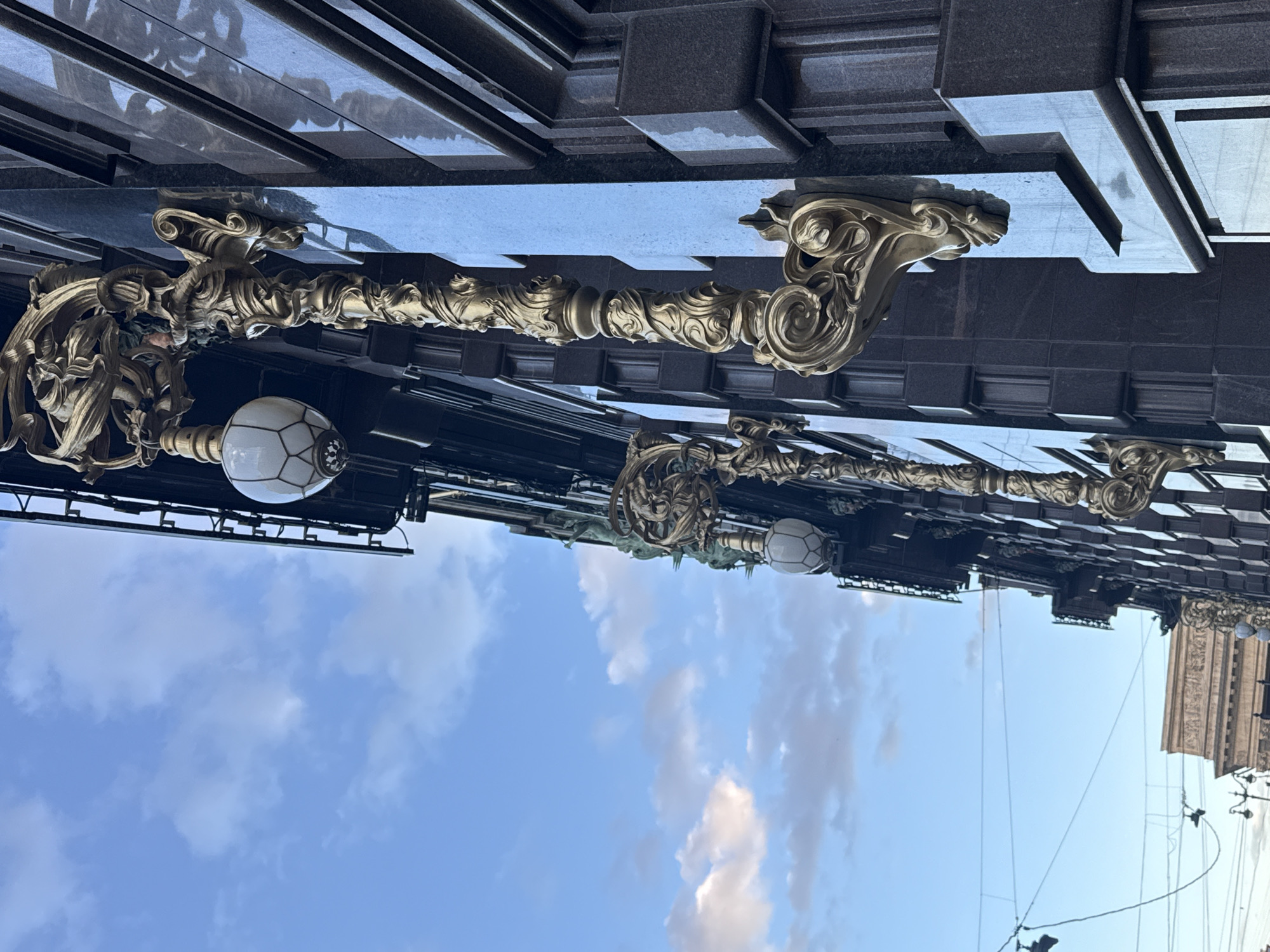 The image shows a close-up, low-angle view of the ornate gold-colored lampposts adorning the facade of the Singer House (Dom Knigi) on Nevsky Prospekt in Saint Petersburg, Russia. The building's dark, granite-like exterior with its distinctive geometric detailing serves as a backdrop. Several lampposts, characterized by their intricate, scrollwork designs and spherical glass globes, are visible along the building's edge. The sky above is a bright blue with scattered white clouds, suggesting it is daytime, likely late afternoon given the soft lighting. In the distance, other architectural elements and what appear to be utility wires are faintly discernible.