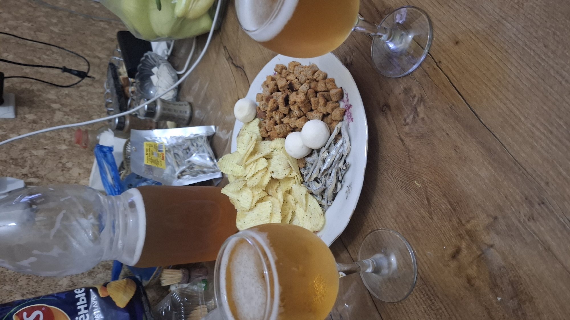 The image displays a close-up of a wooden table featuring a meal and drinks. In the foreground, two wine glasses filled with a golden, bubbly liquid, likely beer, are visible. To the right, a white plate holds a variety of snacks: potato chips, small dried fish, small white spherical objects (possibly cheese or quail eggs), and croutons. Behind the plate, a large glass jug containing the same golden liquid is present, along with a partially visible bag of chips. The background shows a textured wall and various household items, including spice shakers and a plastic bag. The setting appears to be indoors, consistent with a home environment in Shymkent, Kazakhstan. No individuals are depicted in the image.