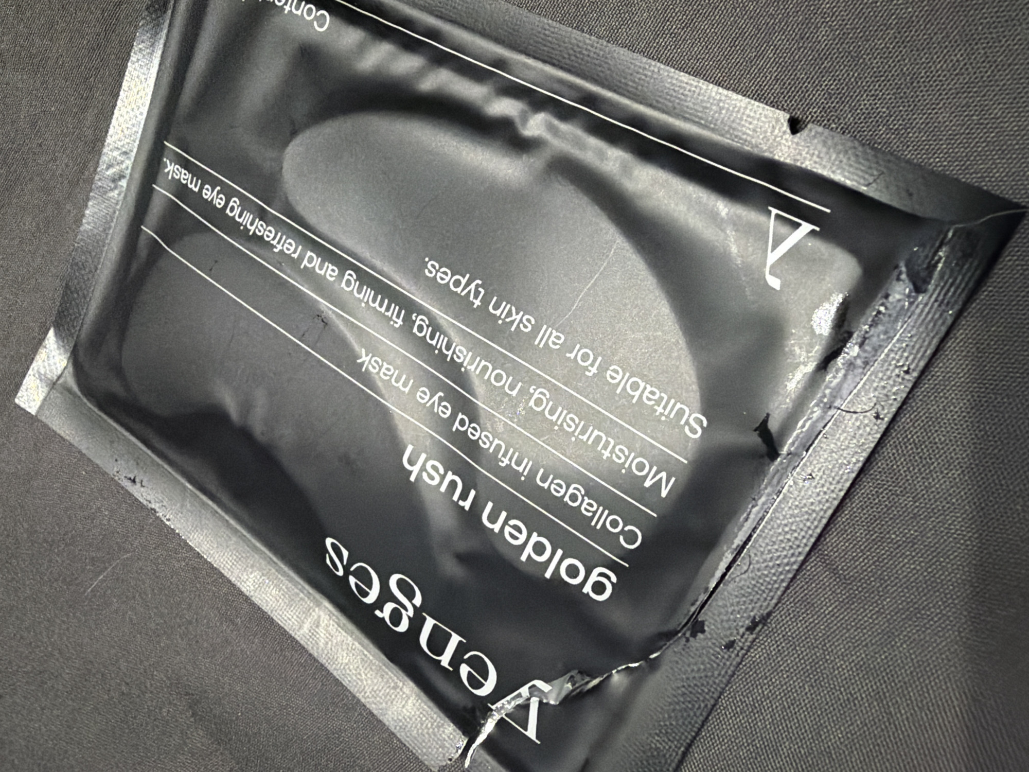 The image presents a close-up, diagonal view of a black, glossy foil packet, likely a single-use sachet for a cosmetic product, resting on a dark grey or black textured fabric surface. The packet exhibits minor creases and reflections, suggesting it's made of a flexible material like plastic or foil.
White text is clearly visible on the packet, identifying the product. The visible text reads: "Venges" (partially visible and inverted, suggesting a brand name), "golden rush" (likely a product line or variant), "Collagen infused eye mask," and a description stating, "Moisturising, nourishing, firming and refreshing eye mask." Additionally, it specifies, "Suitable for all skin types." A truncated word, "Con," appears at the top edge, possibly from "Contents" or "Contains." A stylized white "V" logo is also present on the upper right side of the packet.
The packet shows signs of having been handled or opened. There is a noticeable tear or opening along the bottom-left edge, accompanied by some dark, possibly product-related, residue. The upper-right corner also has a small notch, likely a tear-tab. The background fabric has a fine, uniform weave. The scene depicts a still life of the product, with no human subjects or direct activity taking place. The lighting suggests an indoor setting. There are no visual cues within the image that specifically indicate the location as Burgas, Bulgaria.