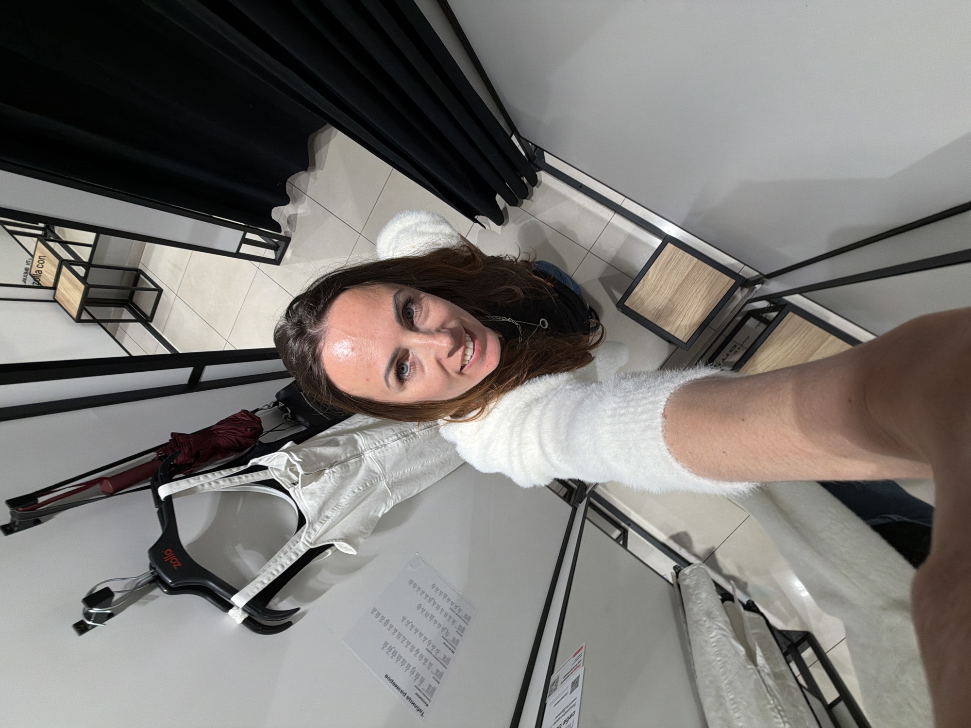 A woman in Birobidzhan, Russia, is taking a selfie in a fitting room. She is wearing a white, fuzzy sweater and has brown hair. The fitting room has a black curtain, tiled floor, shelves, and hanging clothes. A white shirt and a maroon umbrella are visible on a hanger. There are signs on the wall.