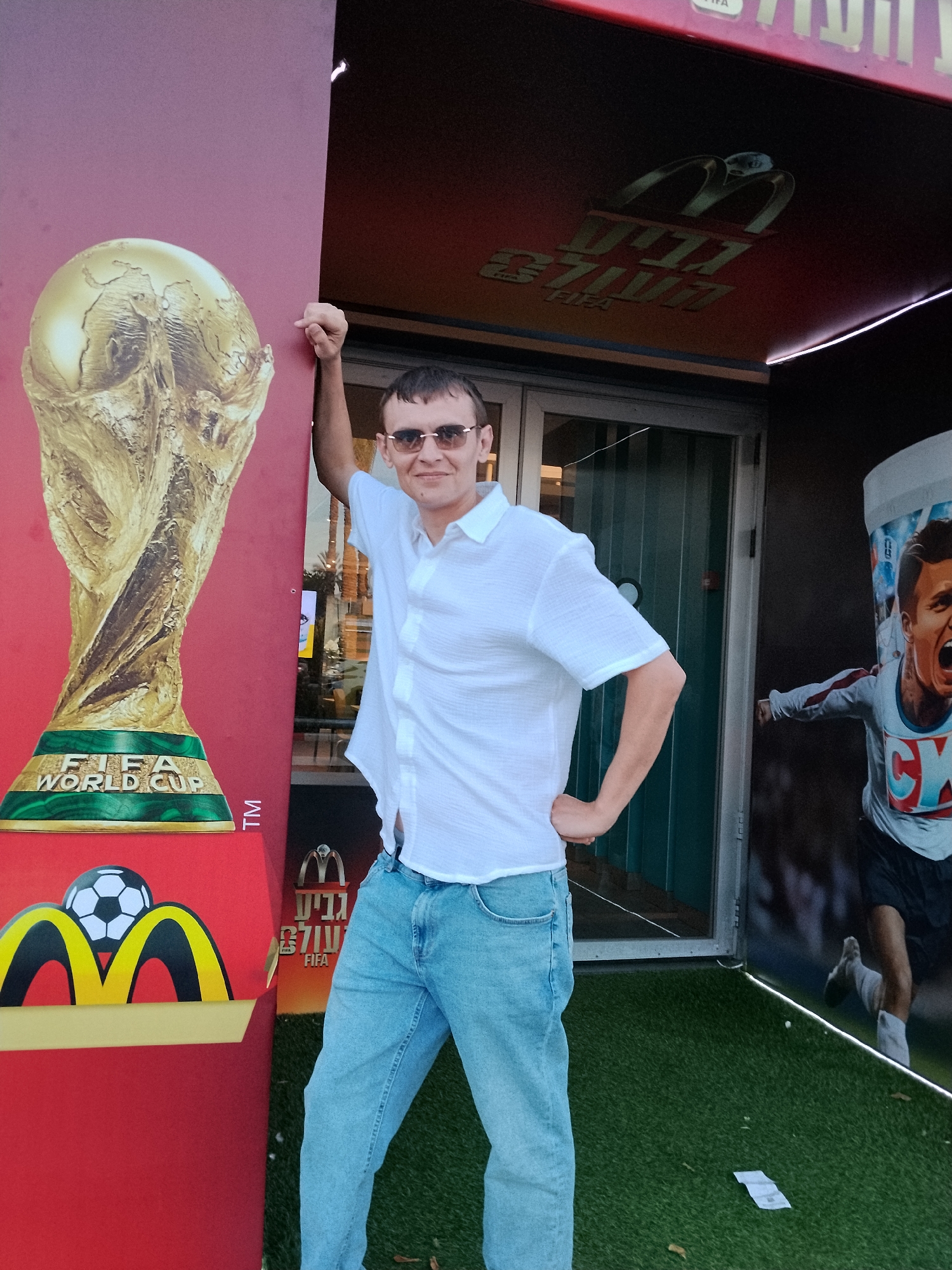 A man is standing in front of a McDonald's kiosk, which is decorated with FIFA World Cup imagery. The kiosk has a large replica of the FIFA World Cup trophy on its left side, and a poster of a soccer player on its right side. The man is wearing a white shirt, blue jeans, and sunglasses. He is smiling and has his hand on his hip. The kiosk is located outdoors, on what appears to be a street or plaza, with artificial grass on the ground. The text on the kiosk is in Hebrew and says "Be'er Sheva, FIFA".